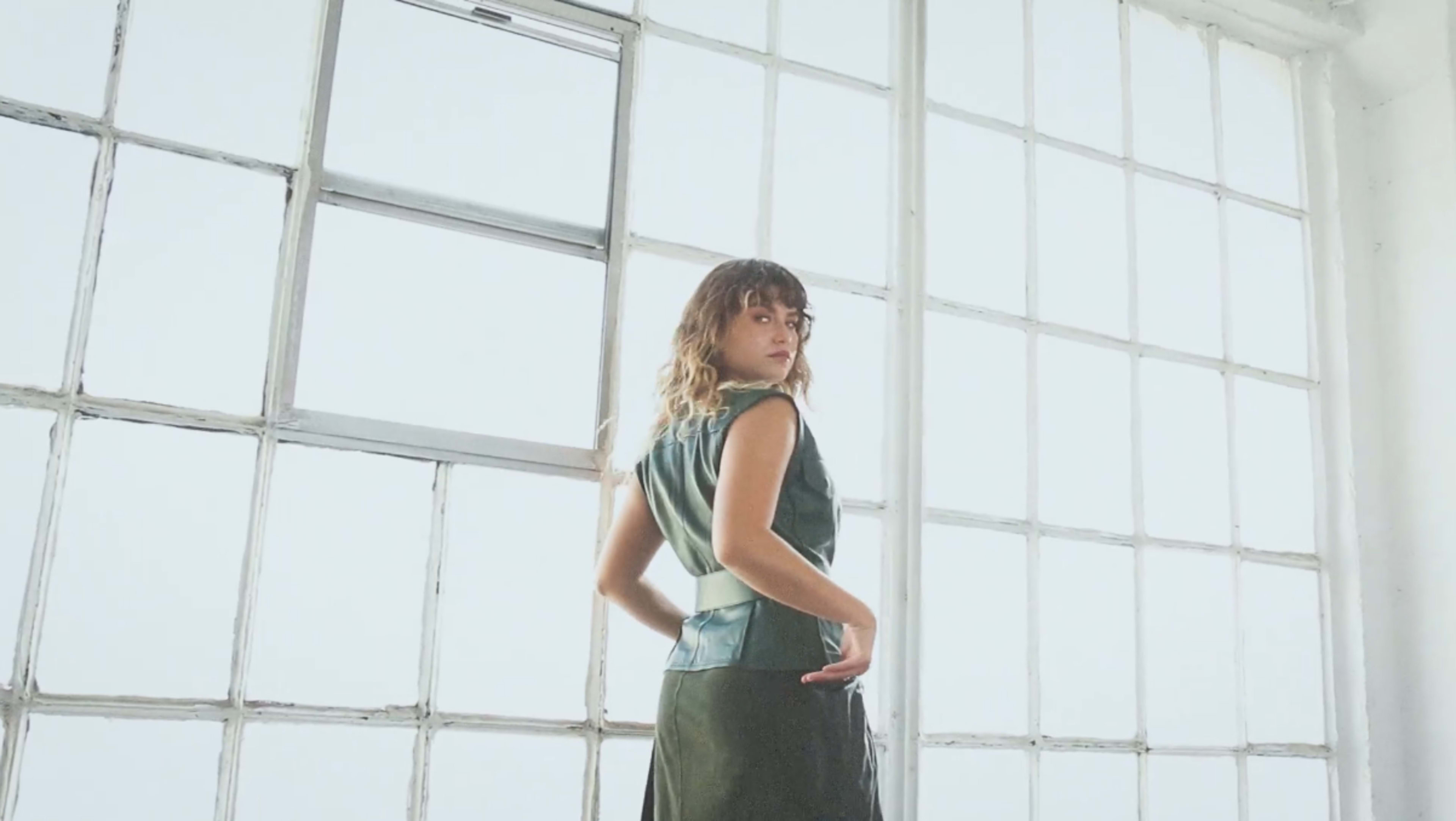 A woman stands facing away from the camera, looking over her shoulder in front of large, industrial-style windows.