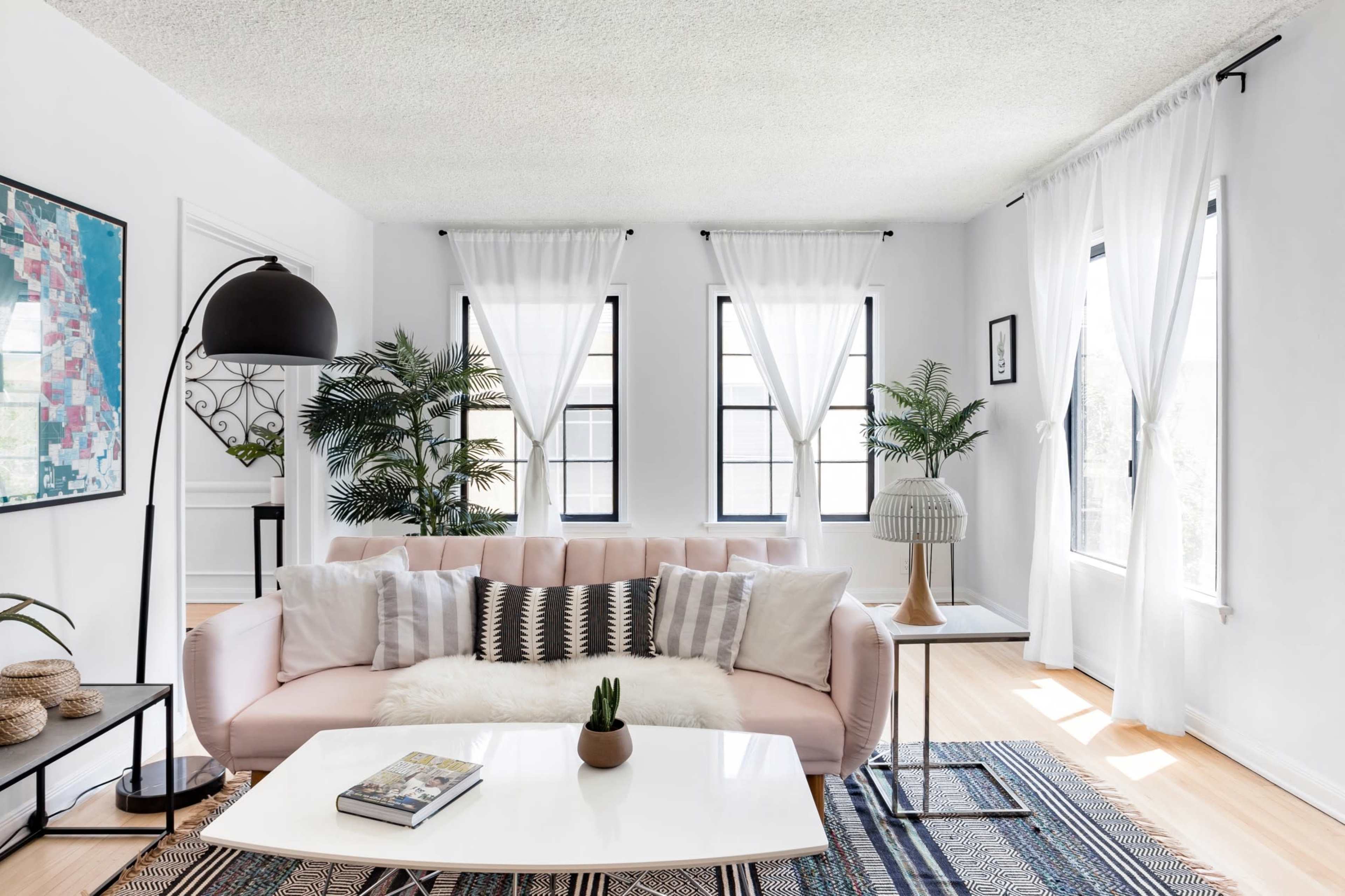The image shows a bright living room featuring a pink sofa, a coffee table, large windows draped with white curtains, and decorative plants.