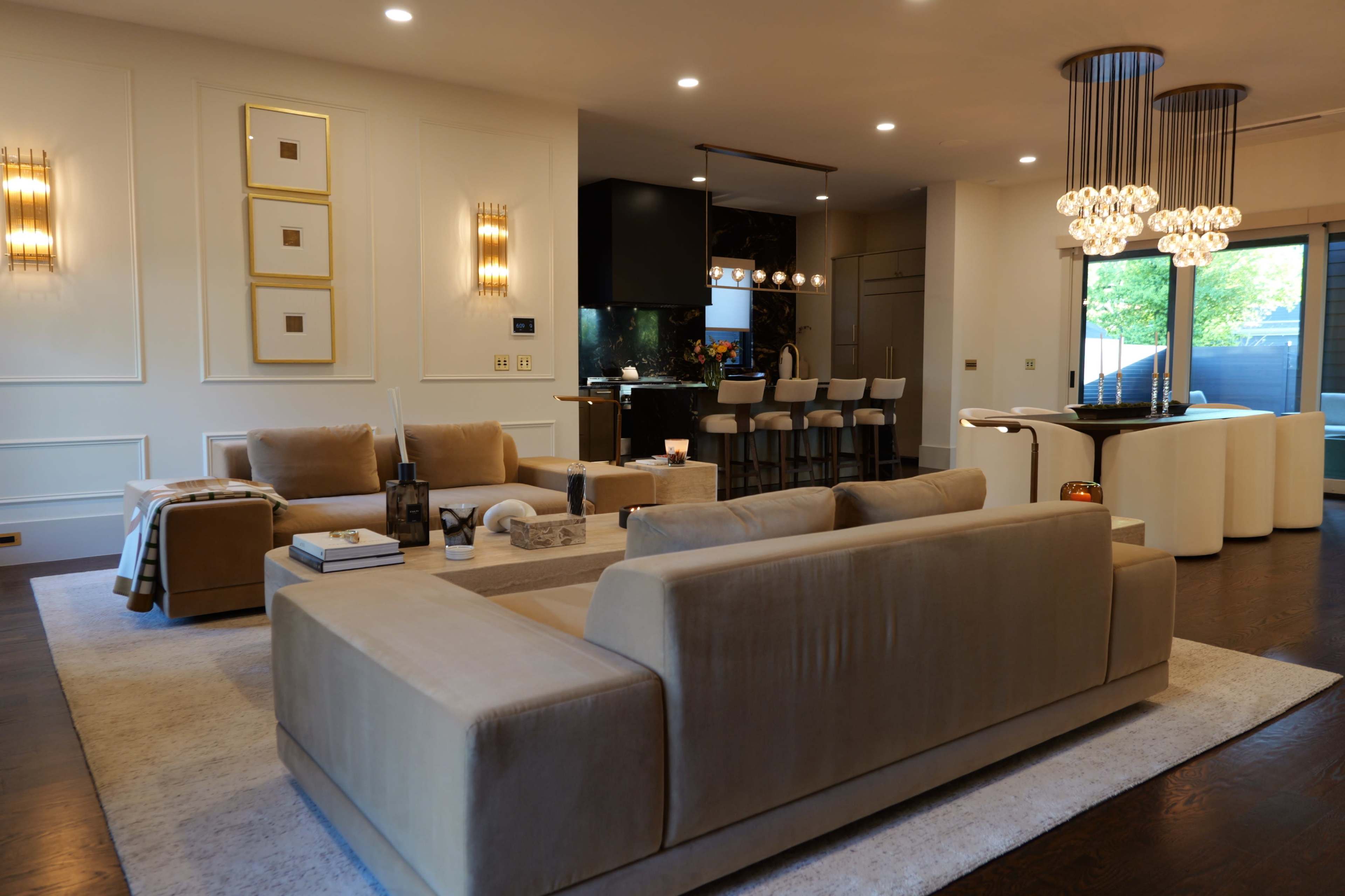 The image shows a modern living room with beige sofas, a coffee table, and a dining area visible in the background, featuring bar stools and contemporary lighting.