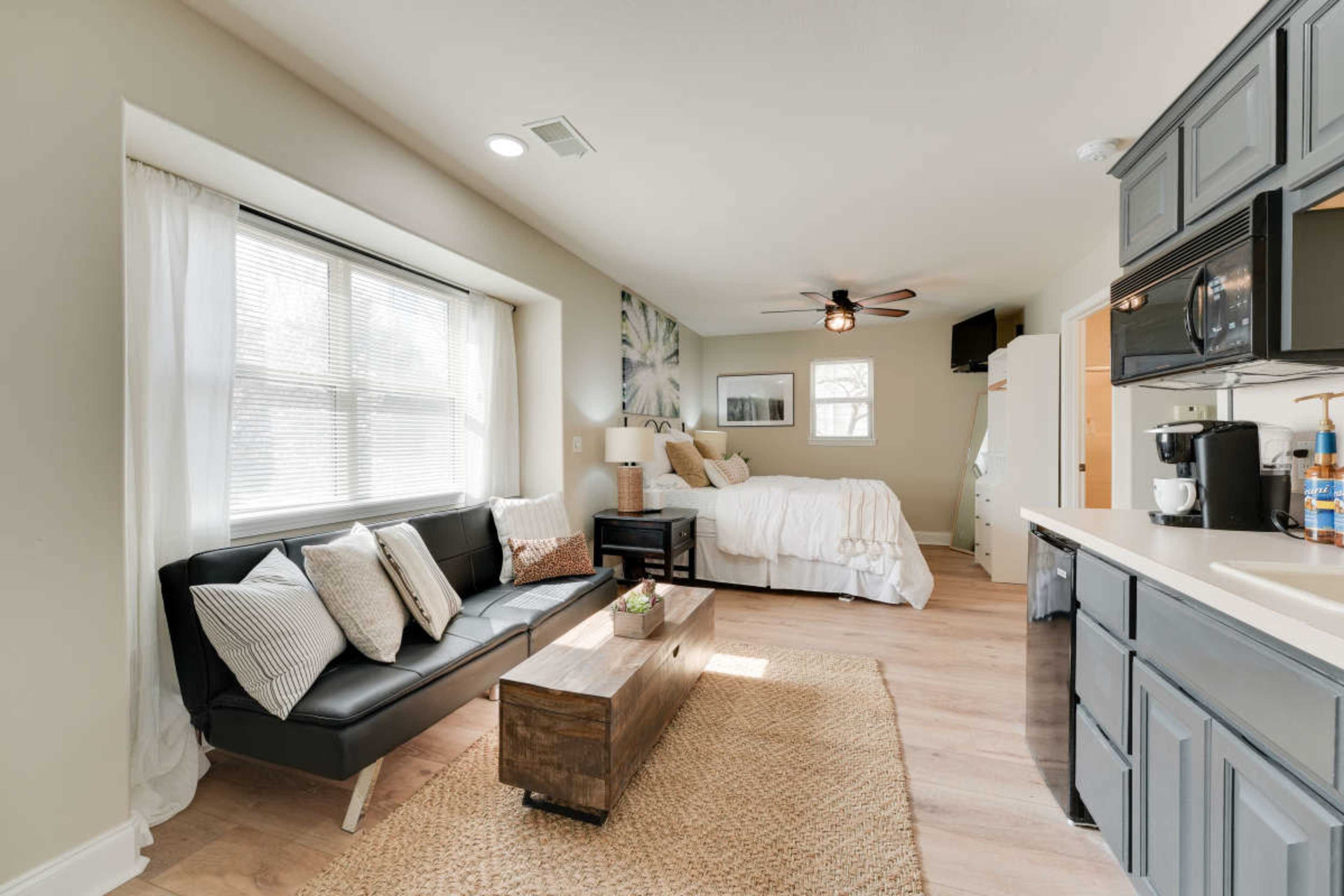 The image shows a compact living space featuring a black sofa, a wooden coffee table, a kitchen area, and a bed in the background.