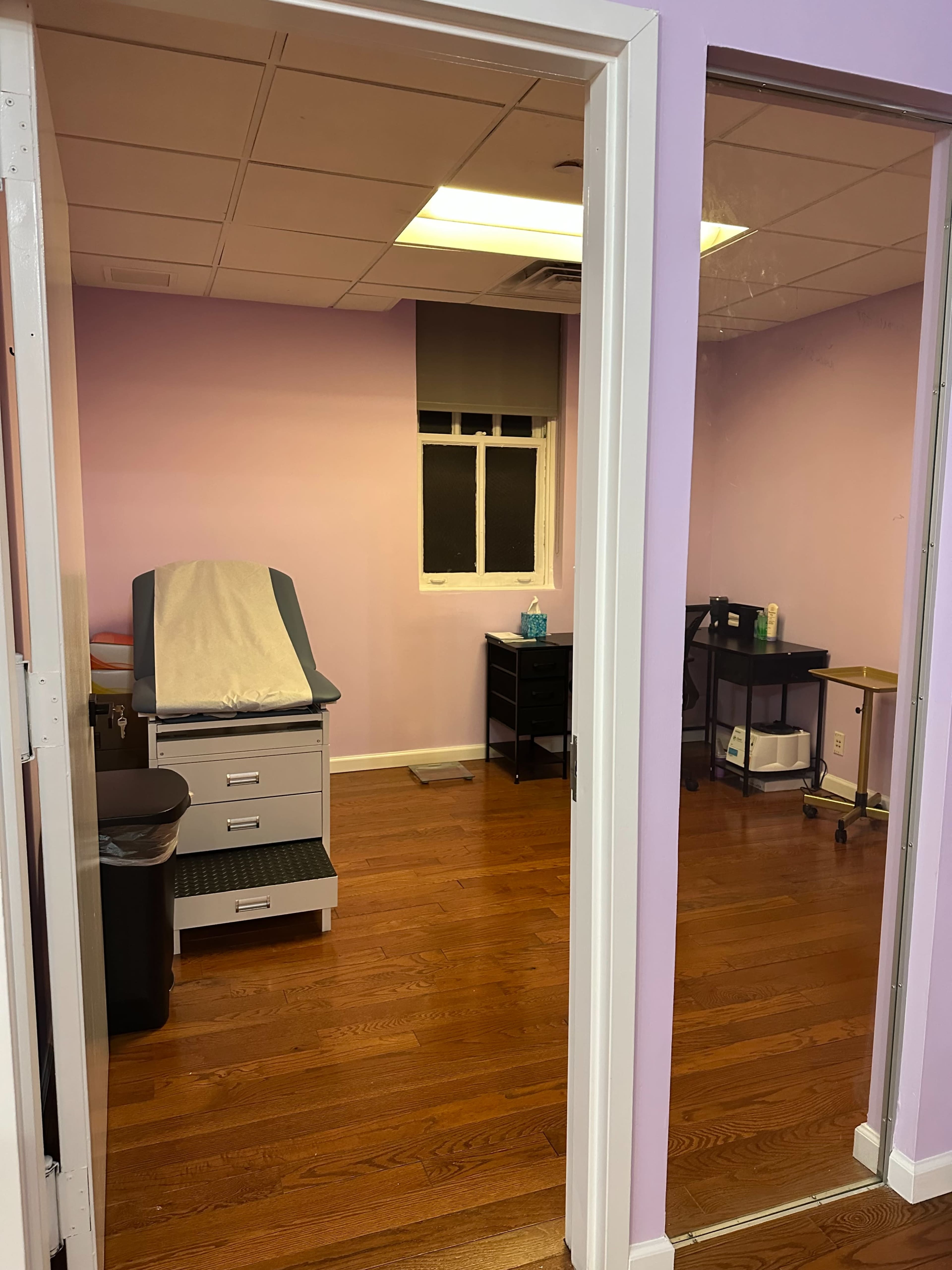 The image shows a medical examination room with a treatment table, trash can, and wooden floors, viewed through a doorway.