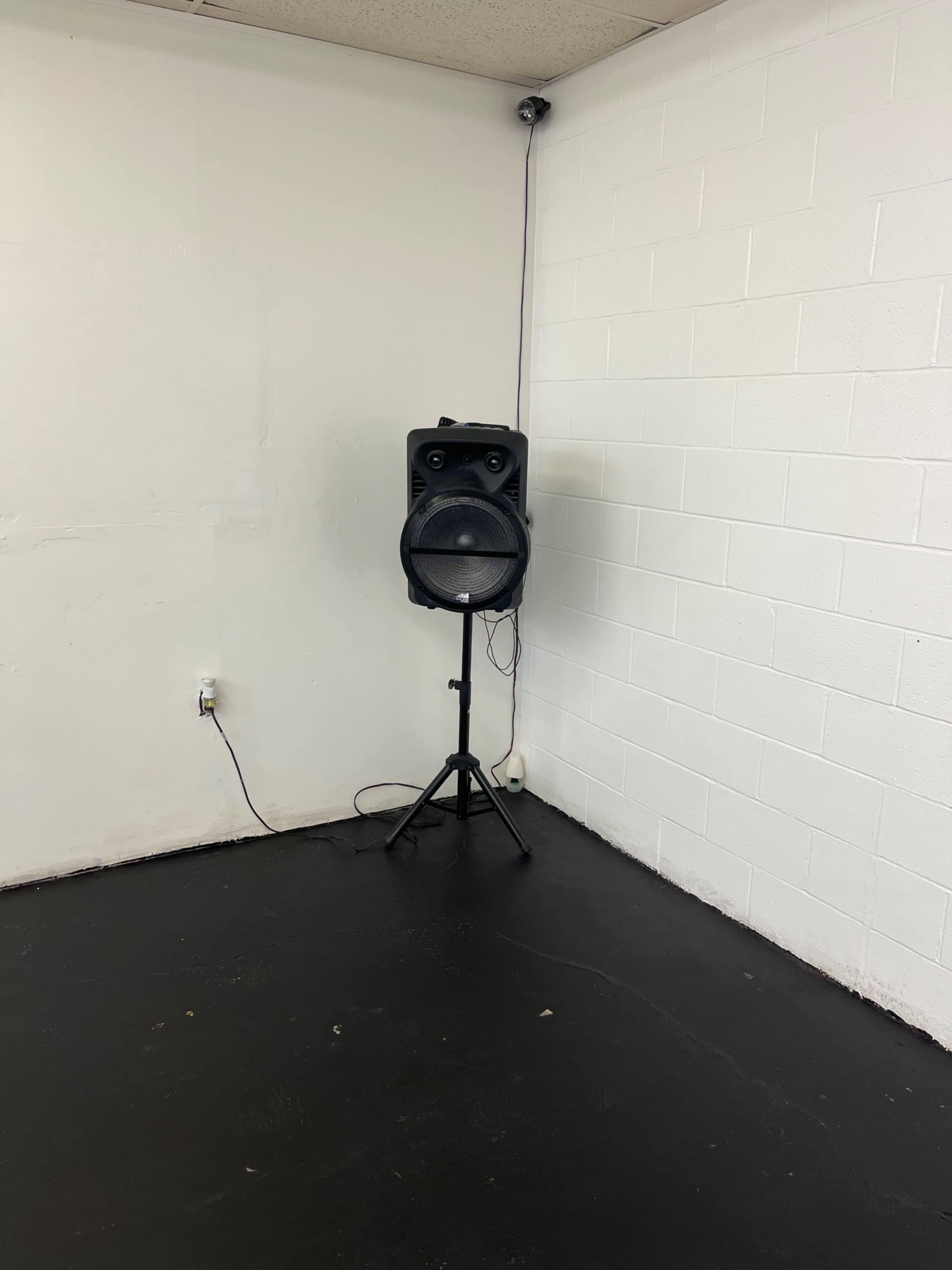 A black speaker on a stand is positioned in the corner of an empty, white-walled room with a dark floor.