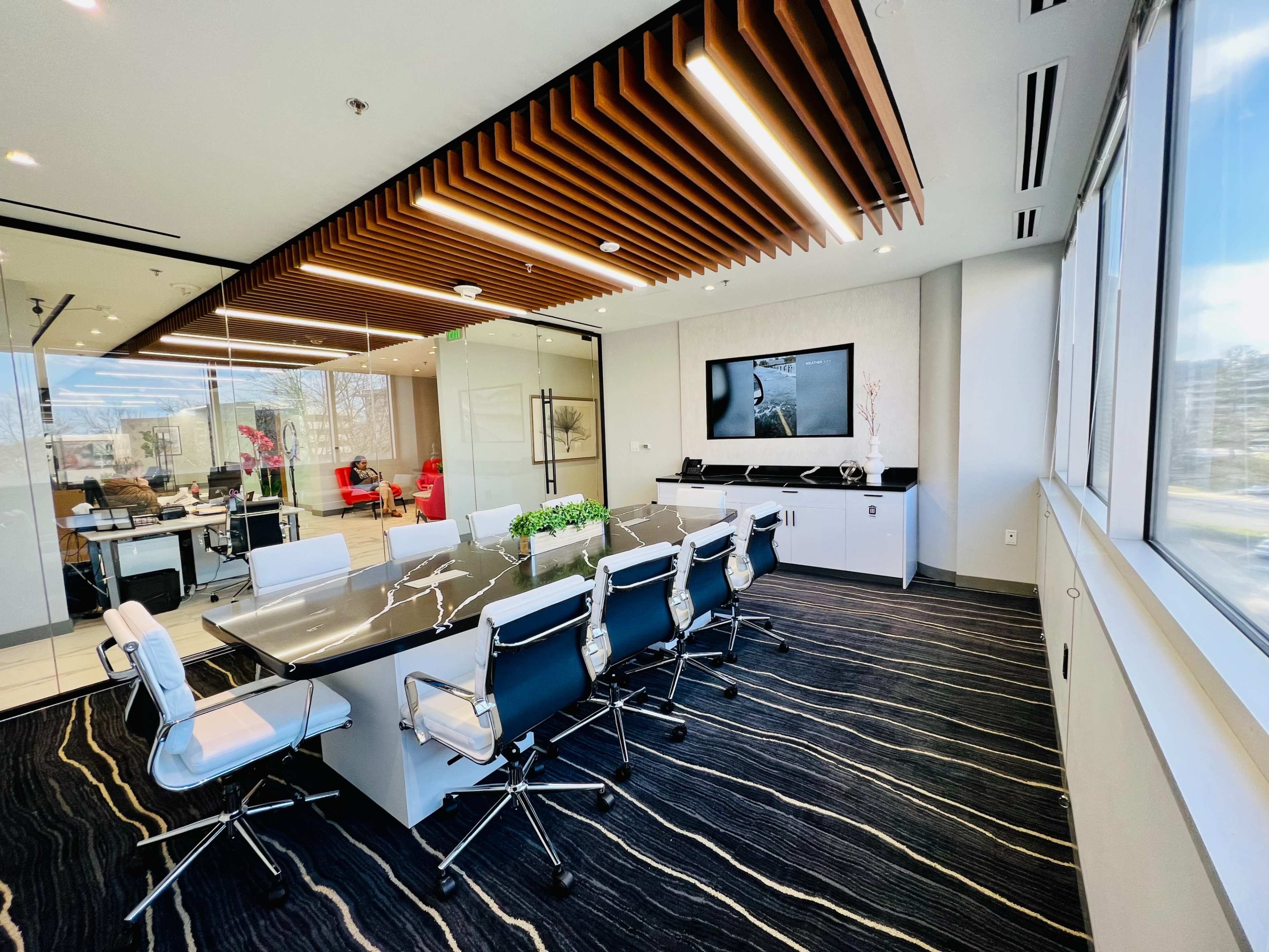 A modern conference room features a large black table surrounded by white and black chairs, with a wall-mounted screen and a view of an adjoining office space.