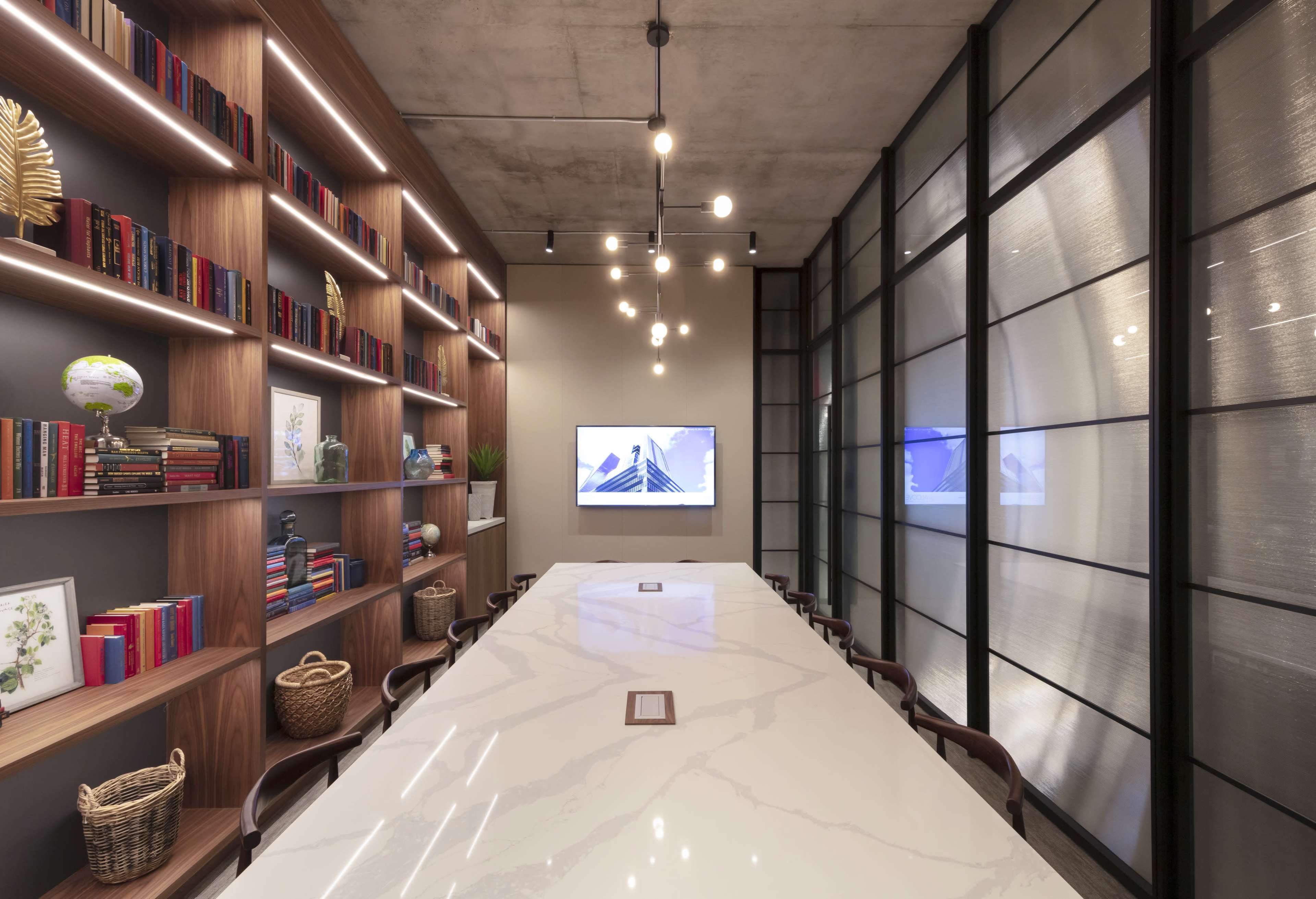 A minimalist conference room features a long marble table, wall-mounted screens, and shelves lined with books and decorative items.