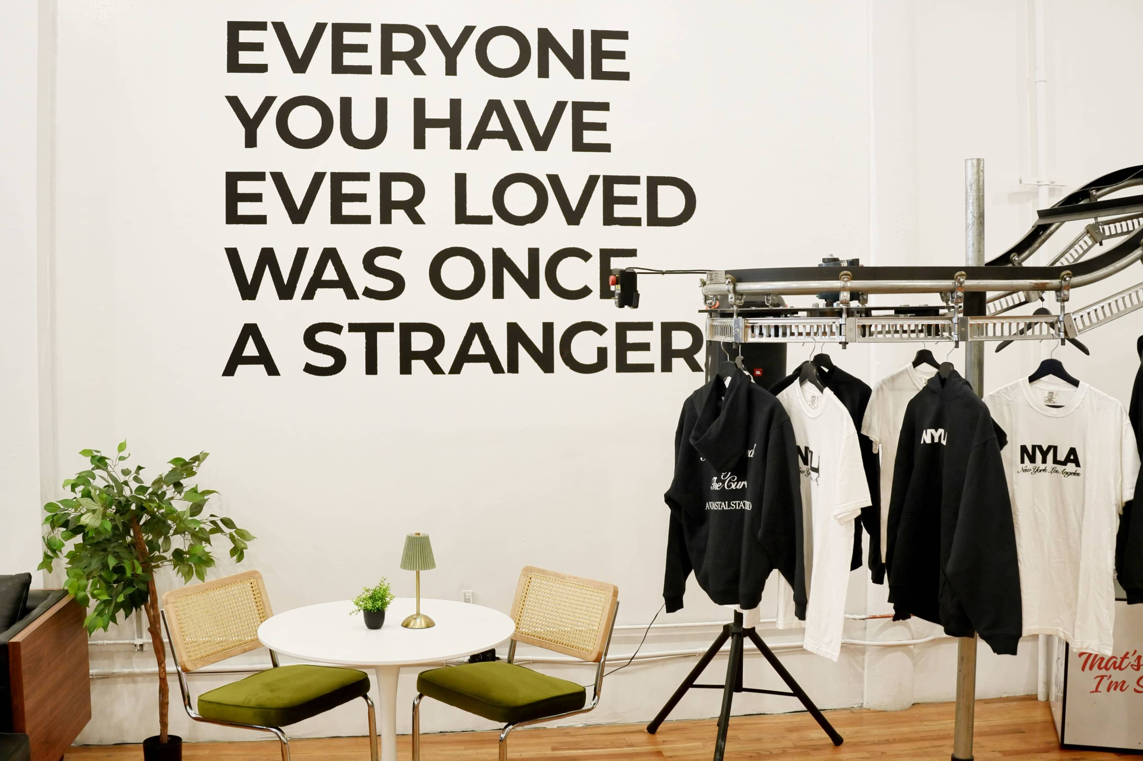 The image shows a minimalist interior space with a wall displaying a large quote, a small round table with chairs, and a clothing rack holding various garments.