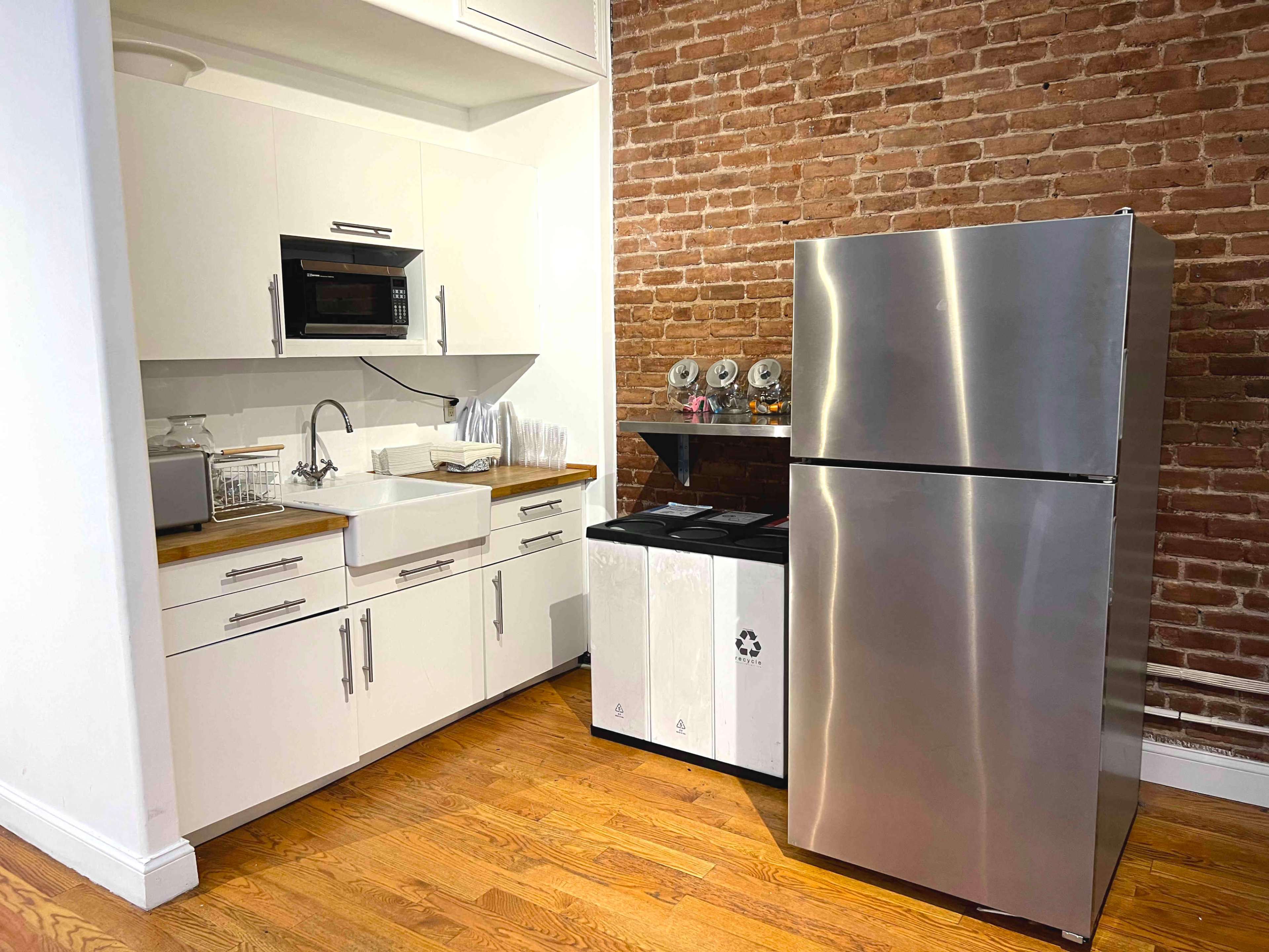 A modern kitchen features a stainless steel refrigerator, a sink with a wooden countertop, and a microwave beside white cabinets against a brick wall.