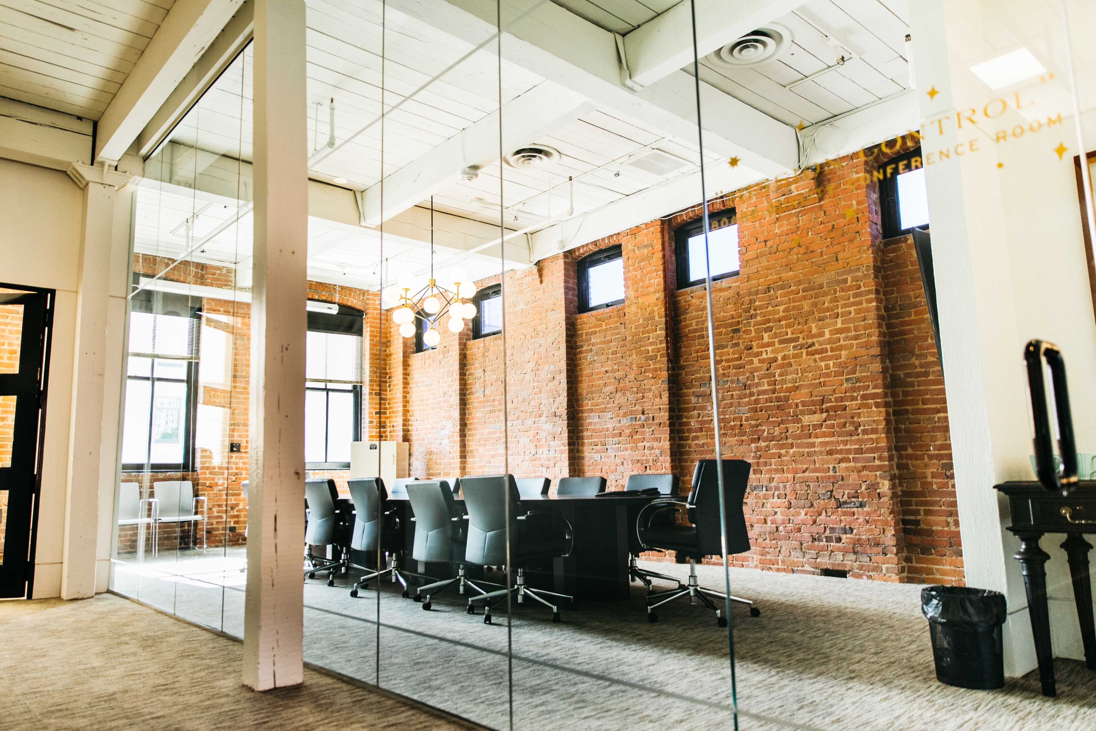 A modern conference room with a long black table and gray chairs is reflected in a glass wall featuring exposed brick.