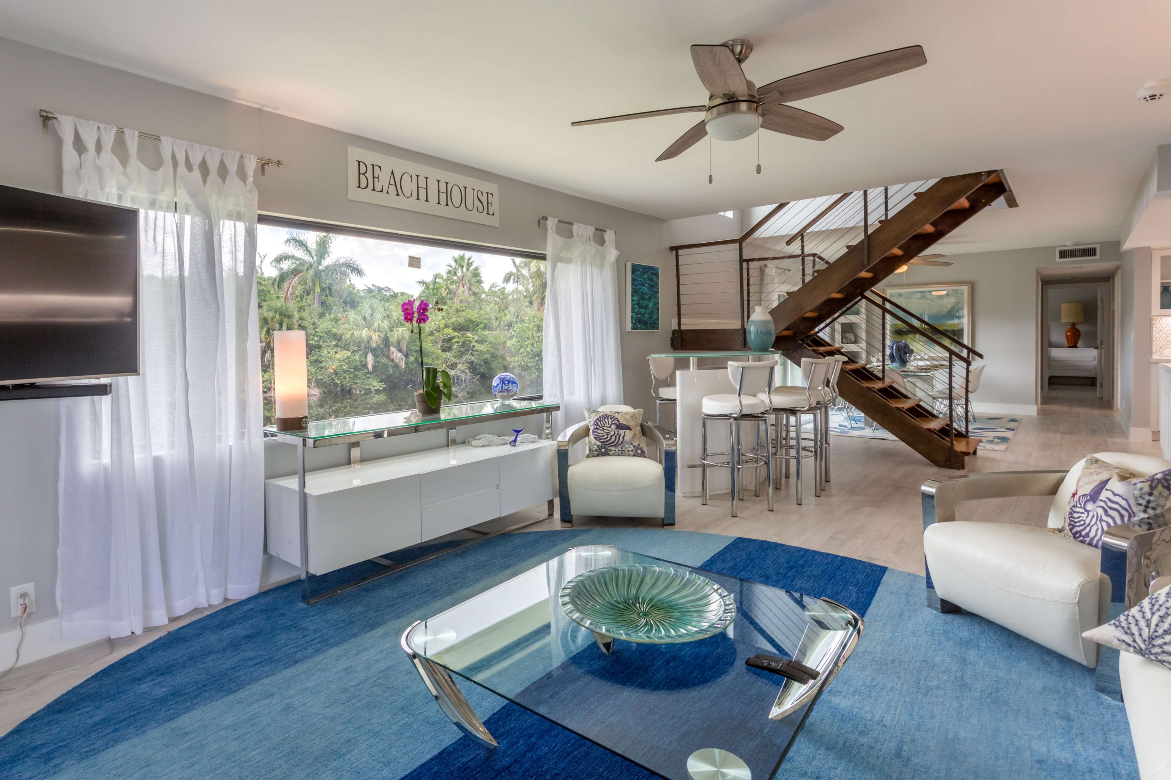 A modern living room with a large window, a blue area rug, a white coffee table, and a staircase leading to an upper level.