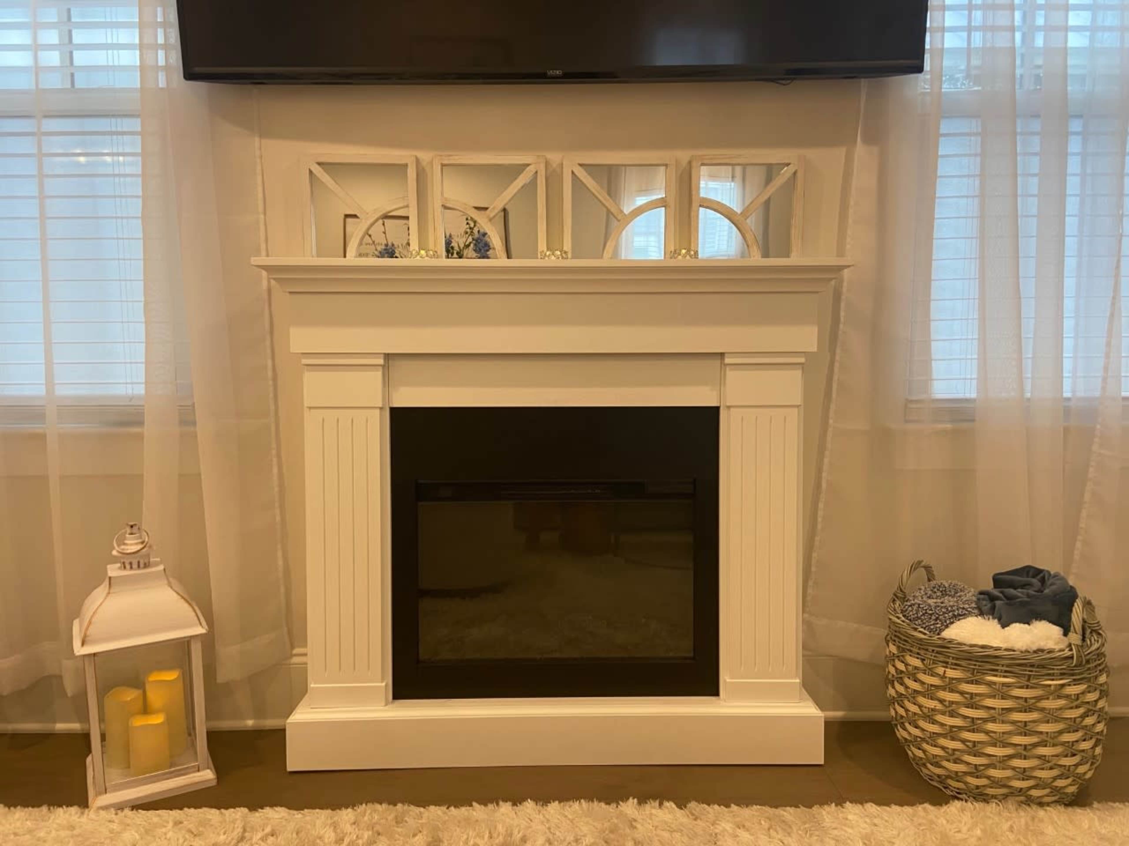A white fireplace with a black insert, surrounded by light curtains and accompanied by a decorative lantern and woven basket.