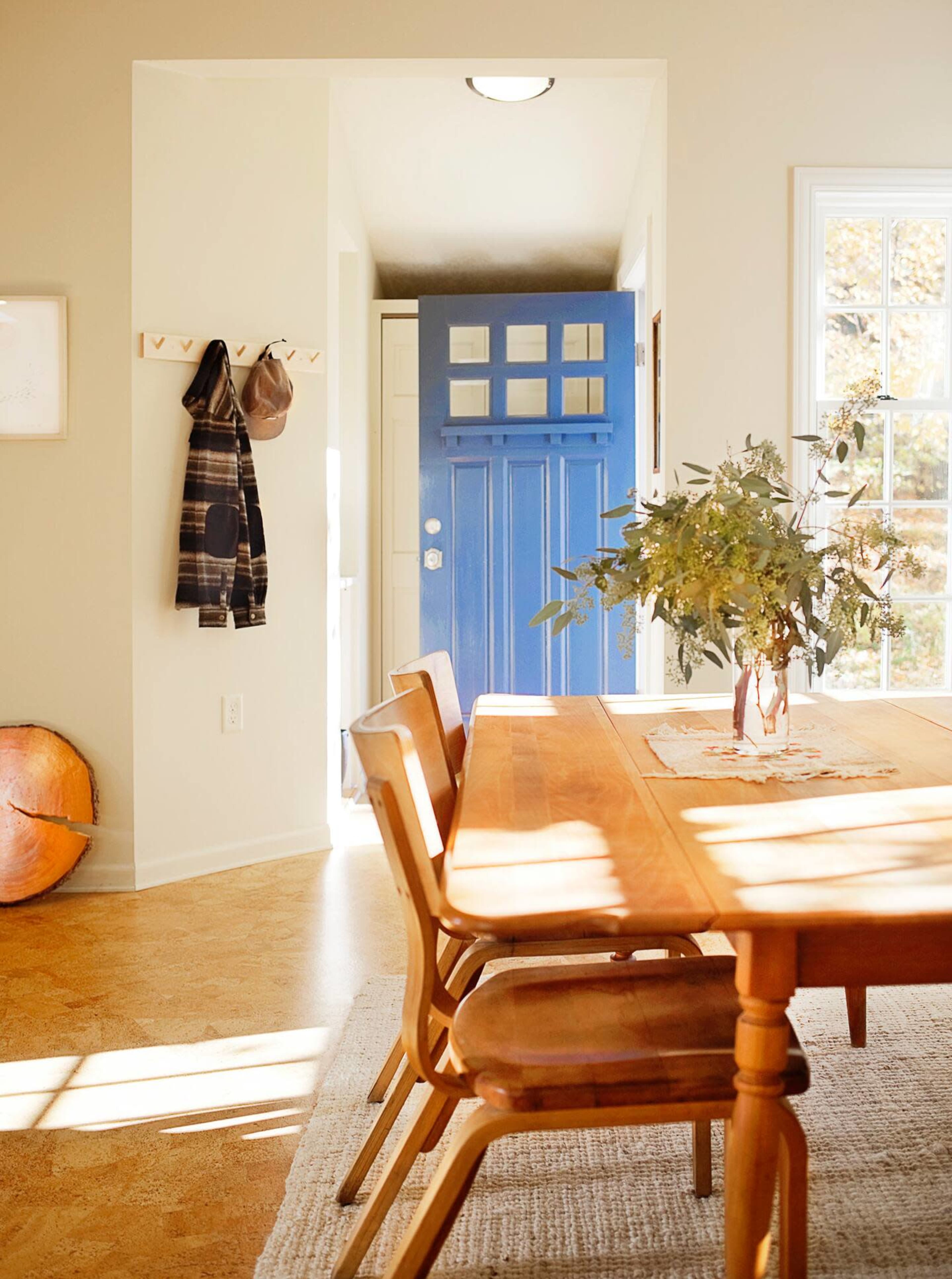 A wooden dining table with chairs is positioned in a brightly lit room, next to a blue front door and a coat rack.