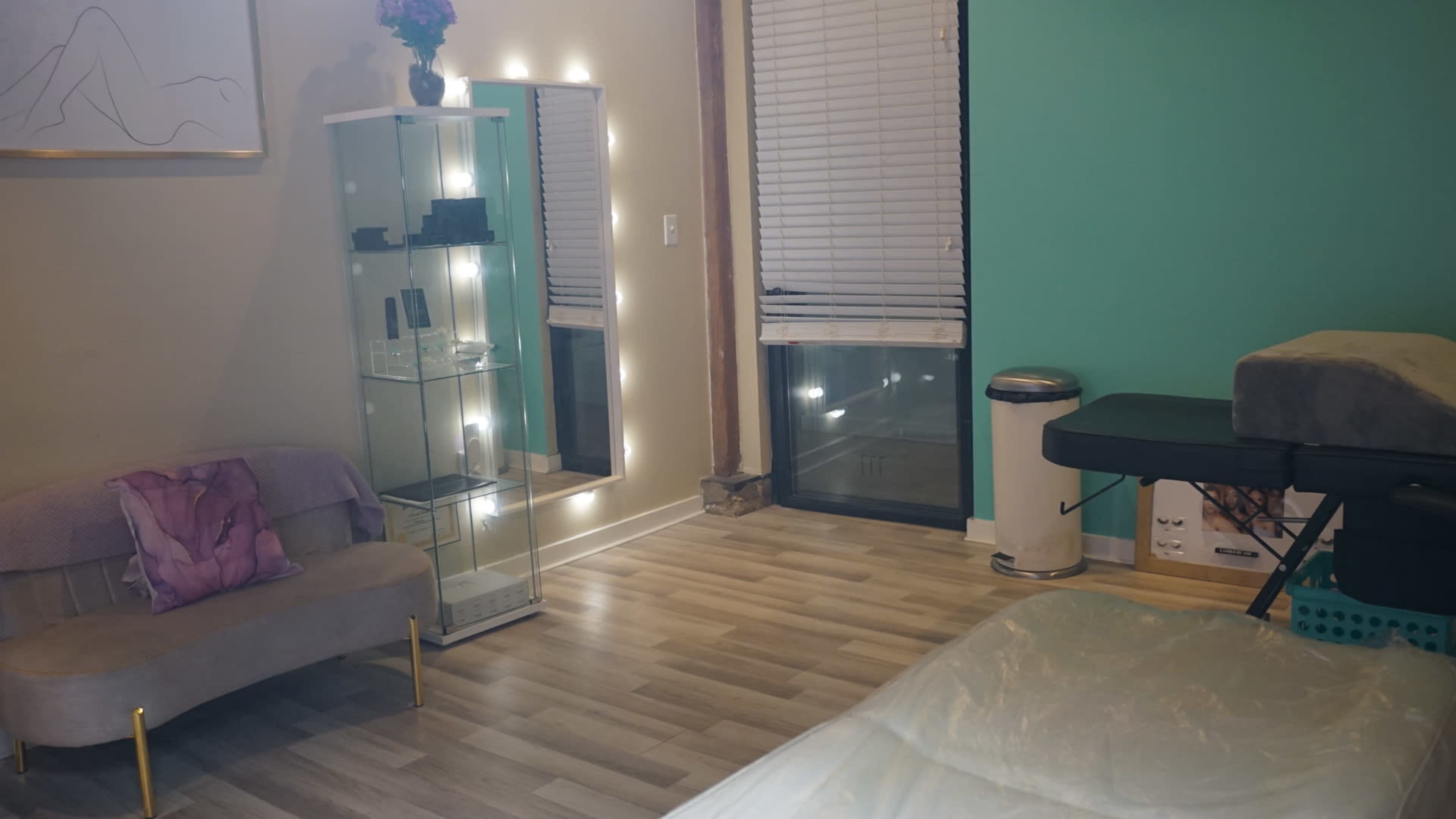 The room features a massage table, a mirror with lights, and a glass display case beside a plush couch against a teal wall.