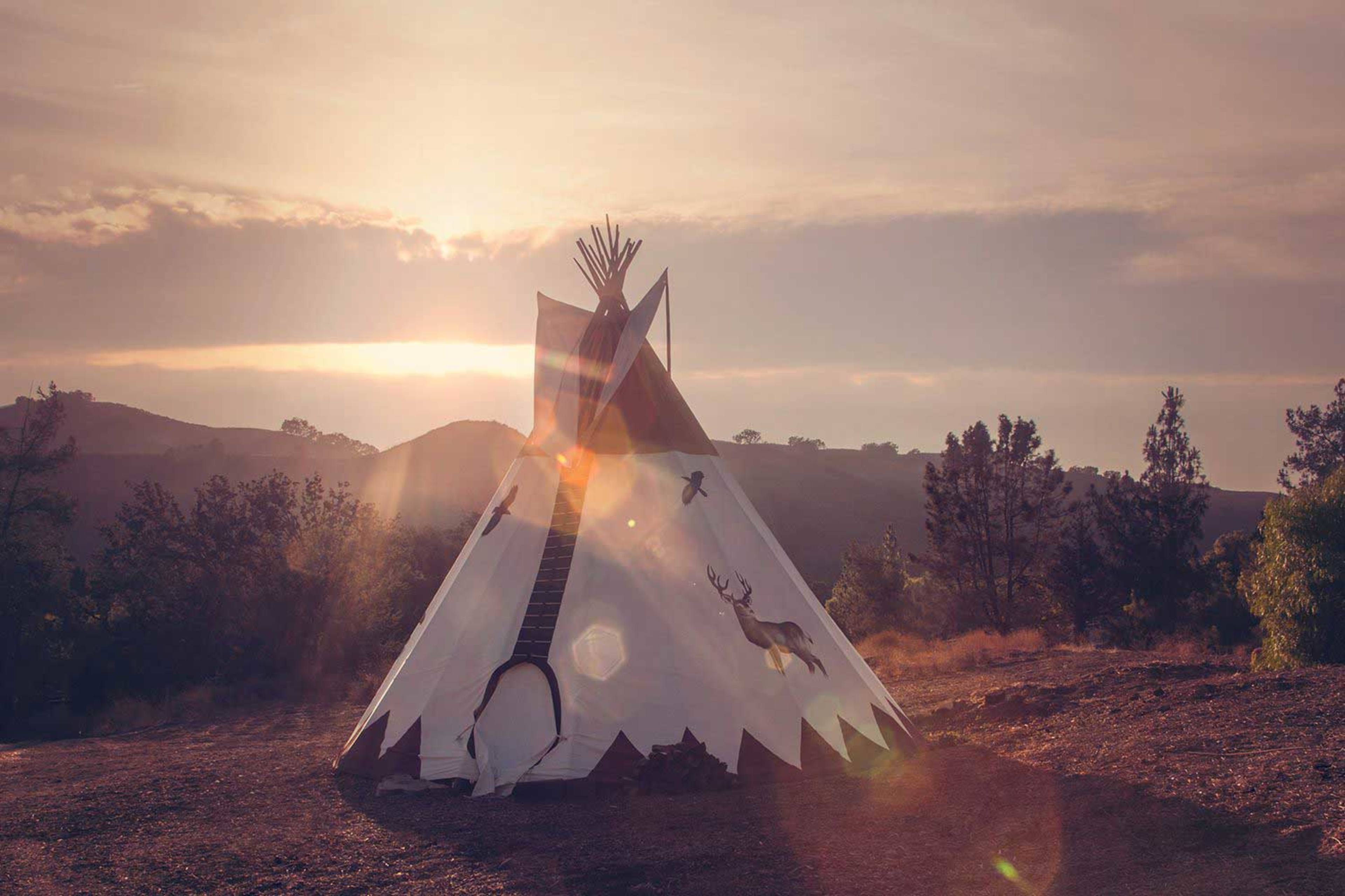 A teepee is set against a backdrop of mountains under a sunset sky.