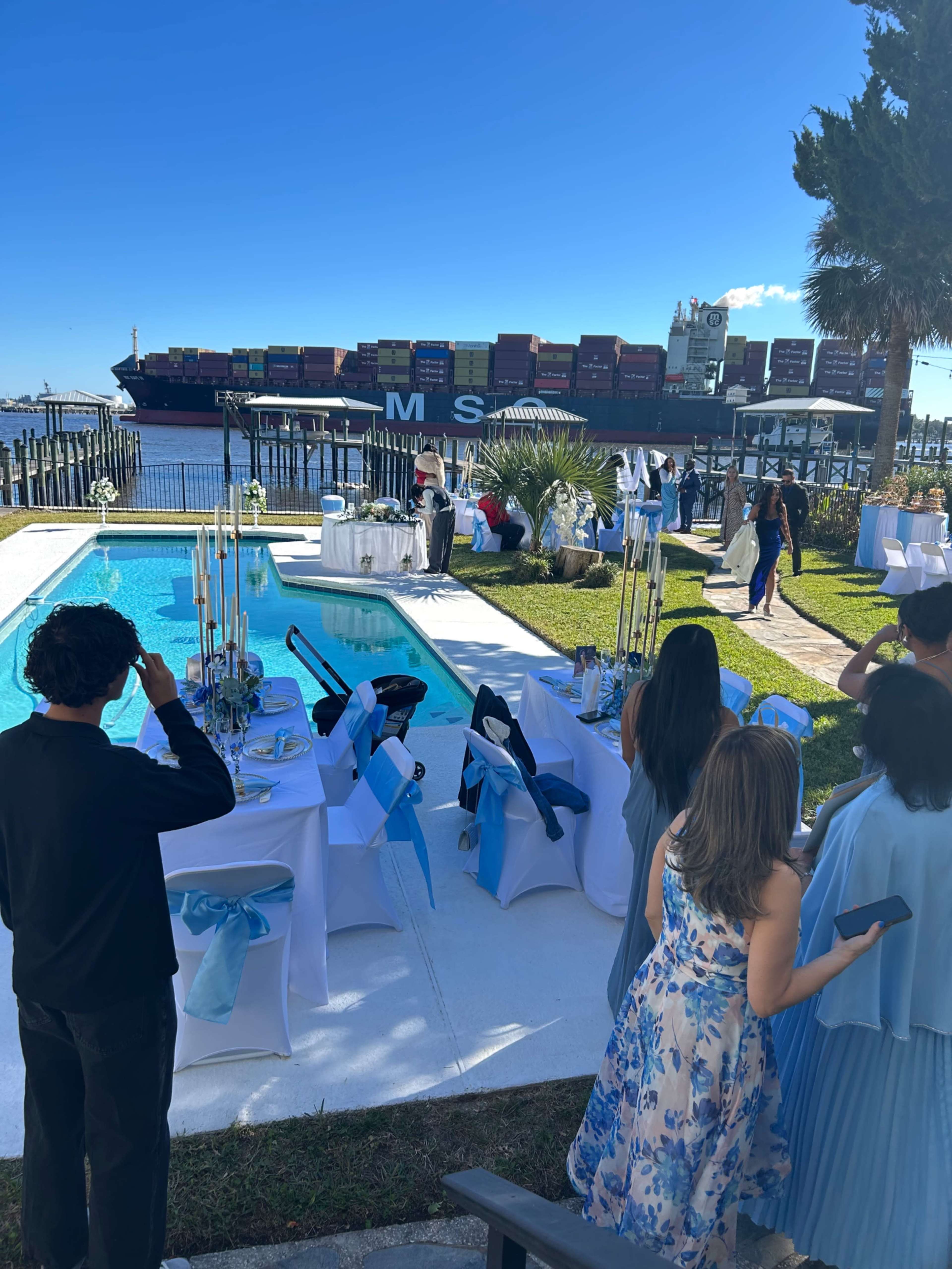 A poolside event is set up with tables and decorations, while a large cargo ship is visible in the background.