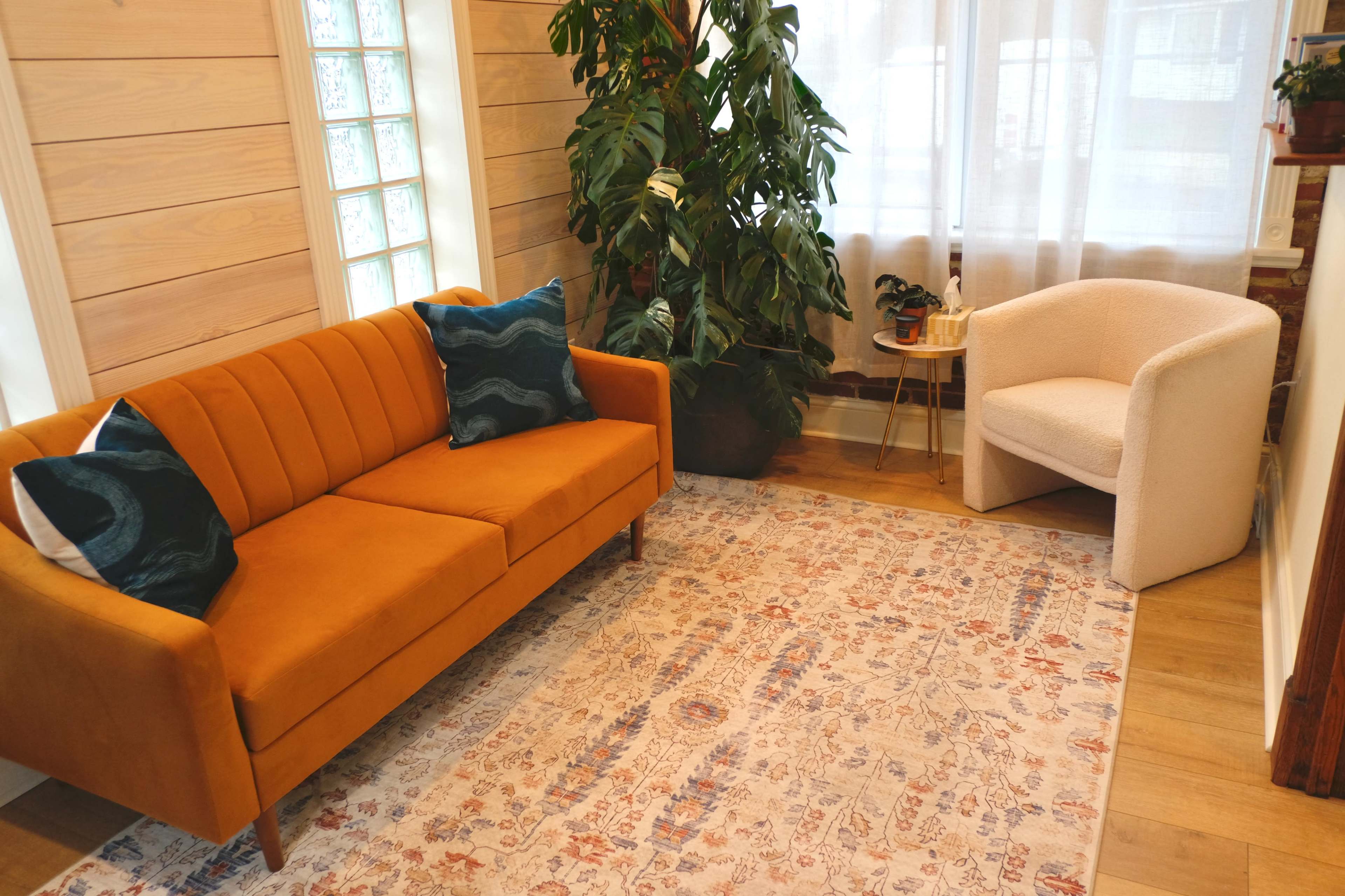 The image shows a cozy interior with an orange sofa and a white chair, accompanied by a leafy plant and a decorative rug on wooden flooring.