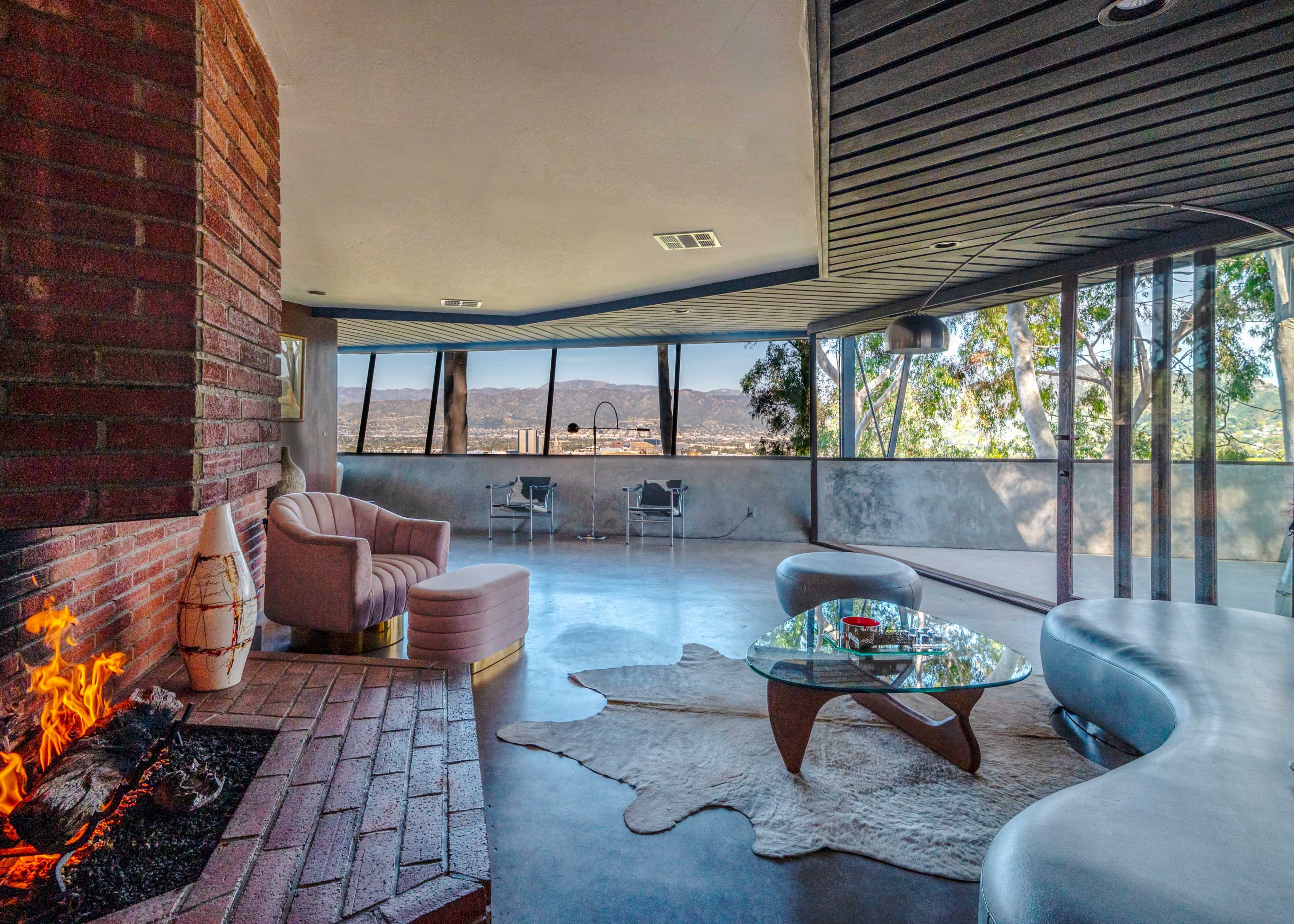 A modern living room features a curved sofa, a glass coffee table, and a fireplace, with large windows offering a view of the surrounding landscape.