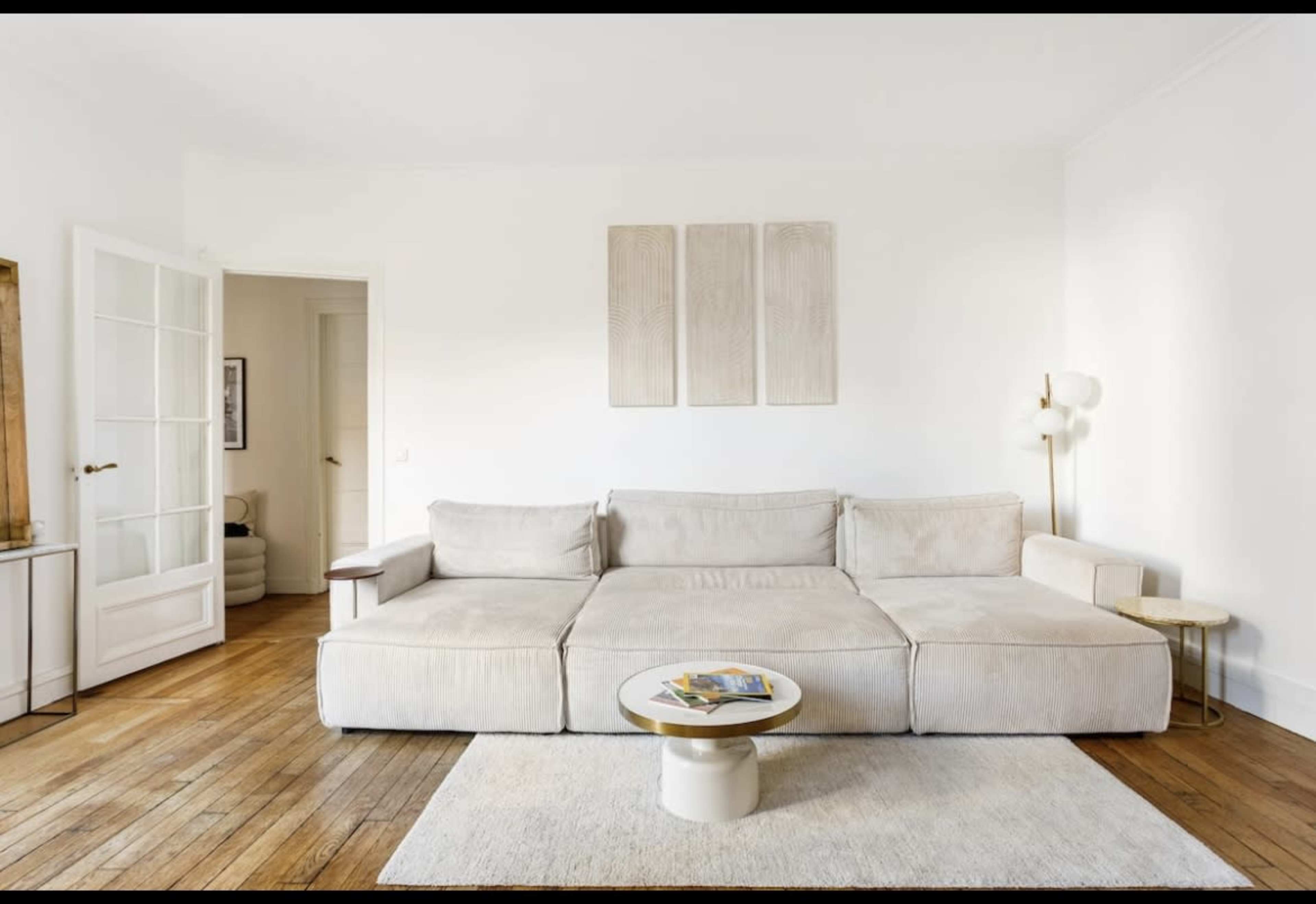 A light-colored living room features a large sectional sofa, a round coffee table, and minimalist wall art above the couch.