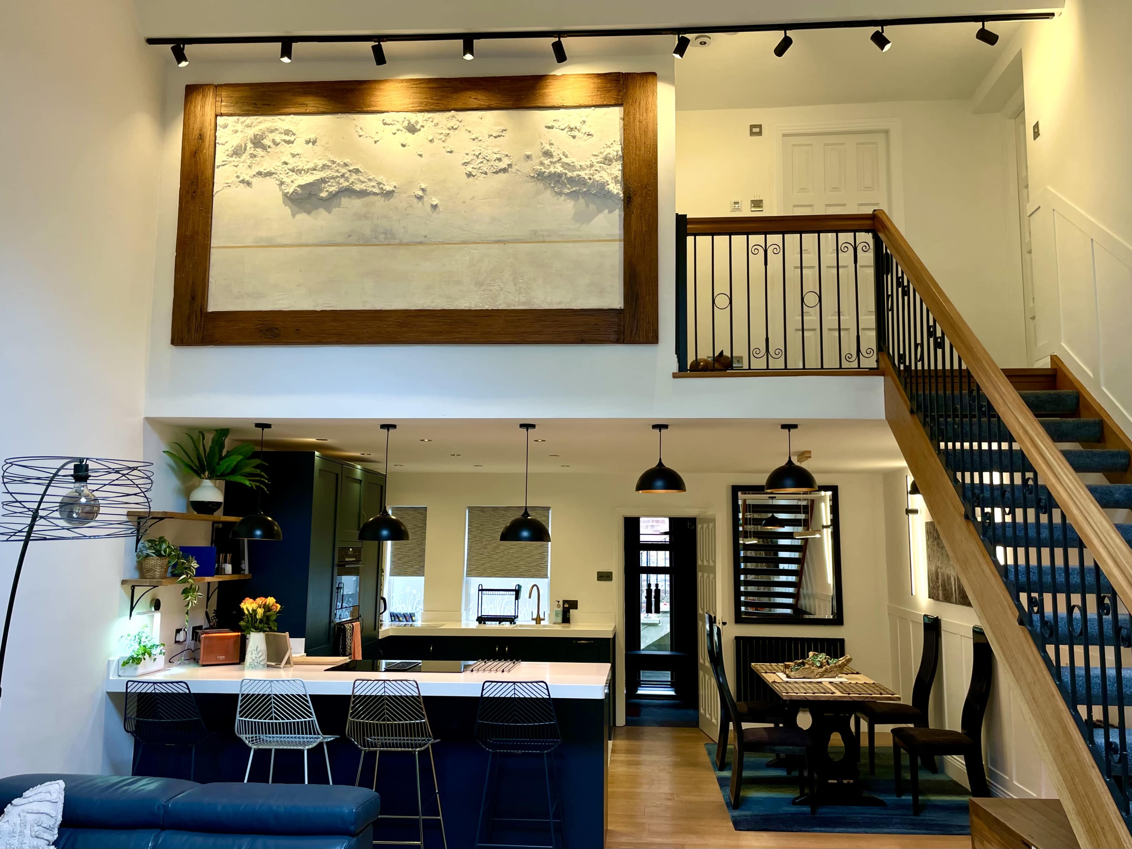 The image shows a modern open-concept kitchen and living room with a large artwork above the dining area and stairs leading to a balcony.