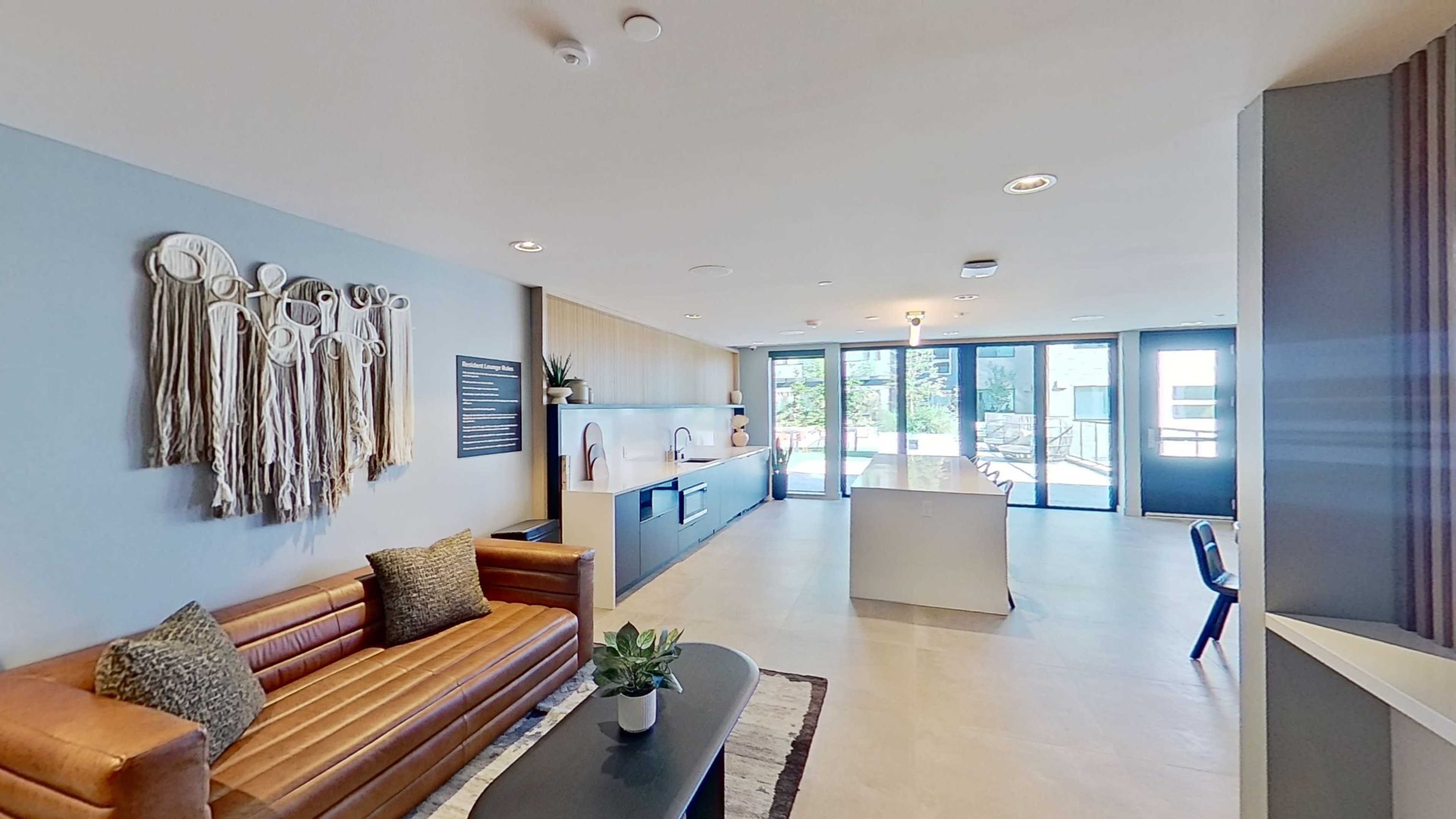 The image shows a modern lounge area featuring a brown leather sofa, a coffee table, and an open kitchen space with large windows that allow natural light to fill the room.