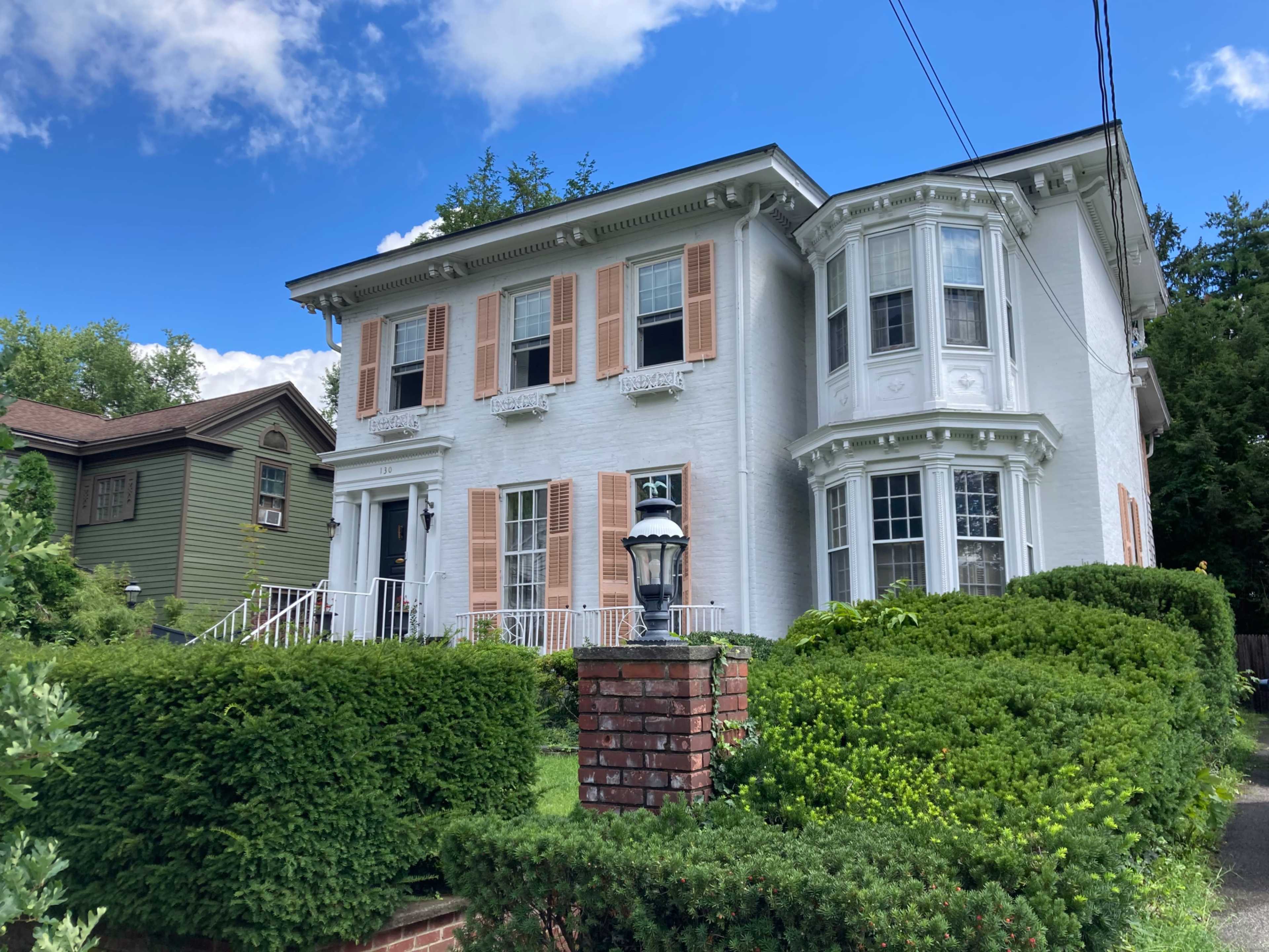 A large white historic house with orange shutters and a round bay window, surrounded by neatly trimmed green bushes and a brick fence.