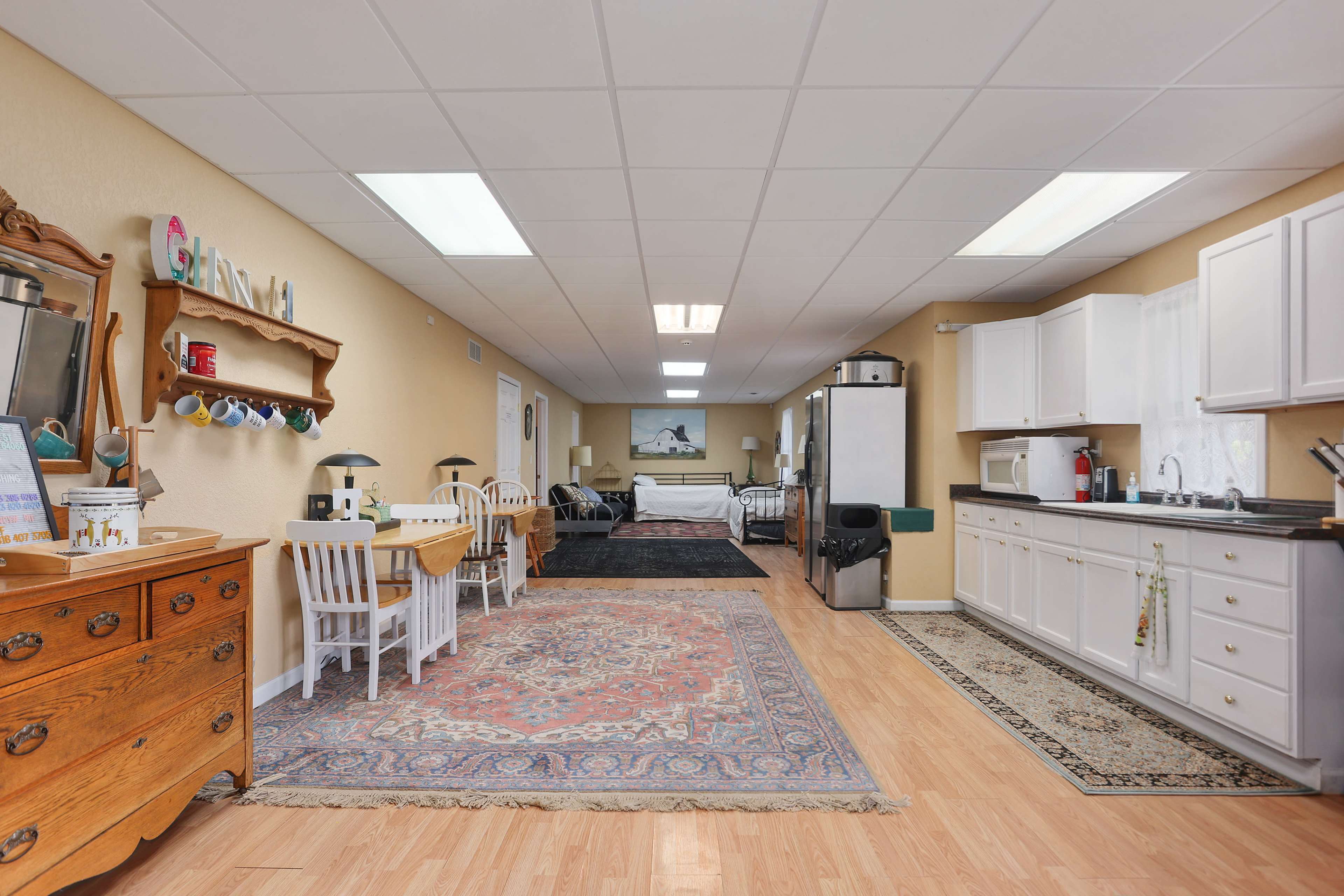 The image shows a spacious room featuring a kitchen area with white cabinets, a dining table with white chairs, and a carpeted living space at the far end.