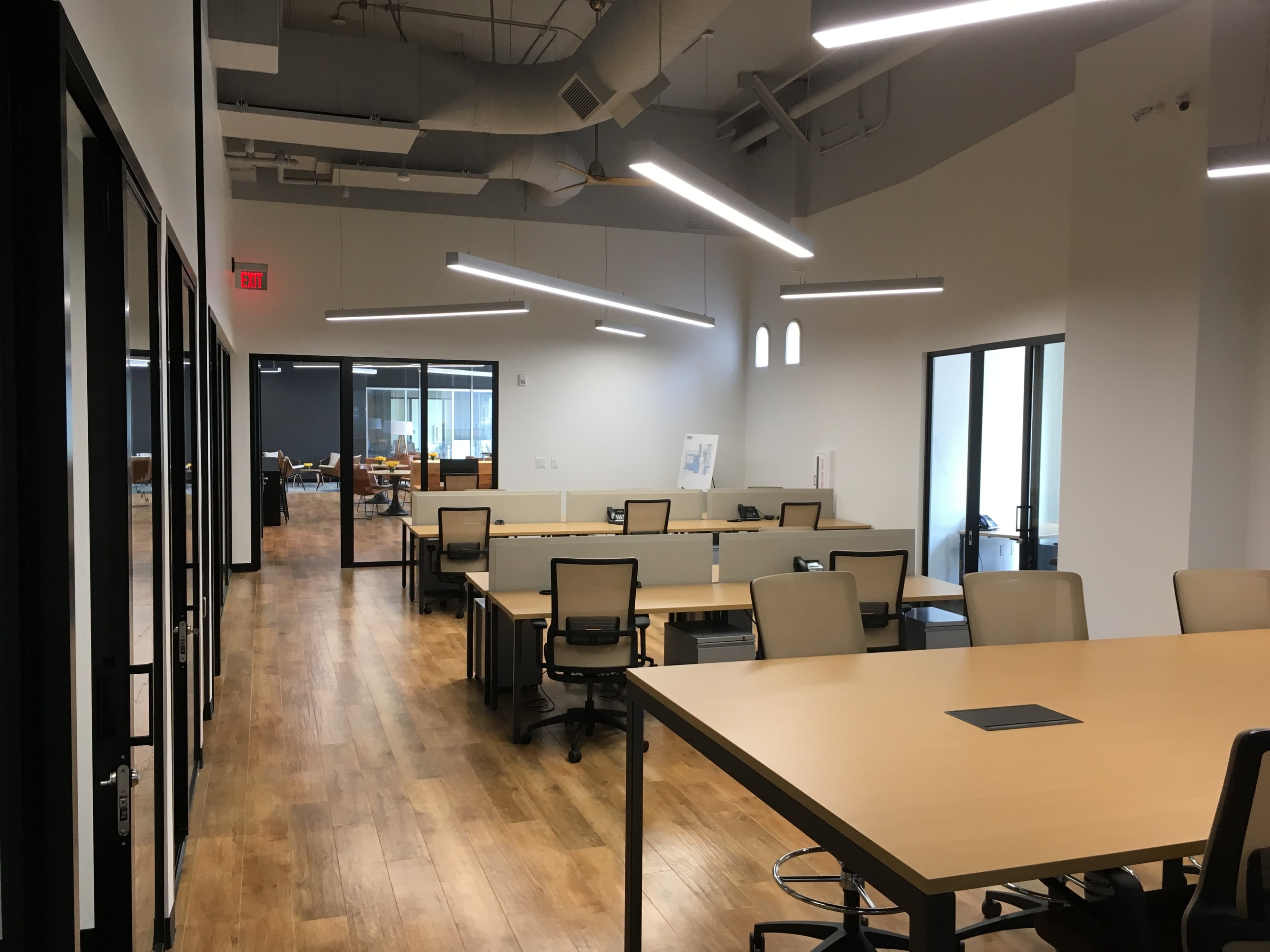 A modern office space with multiple desks arranged in a row, featuring large windows and overhead lighting.
