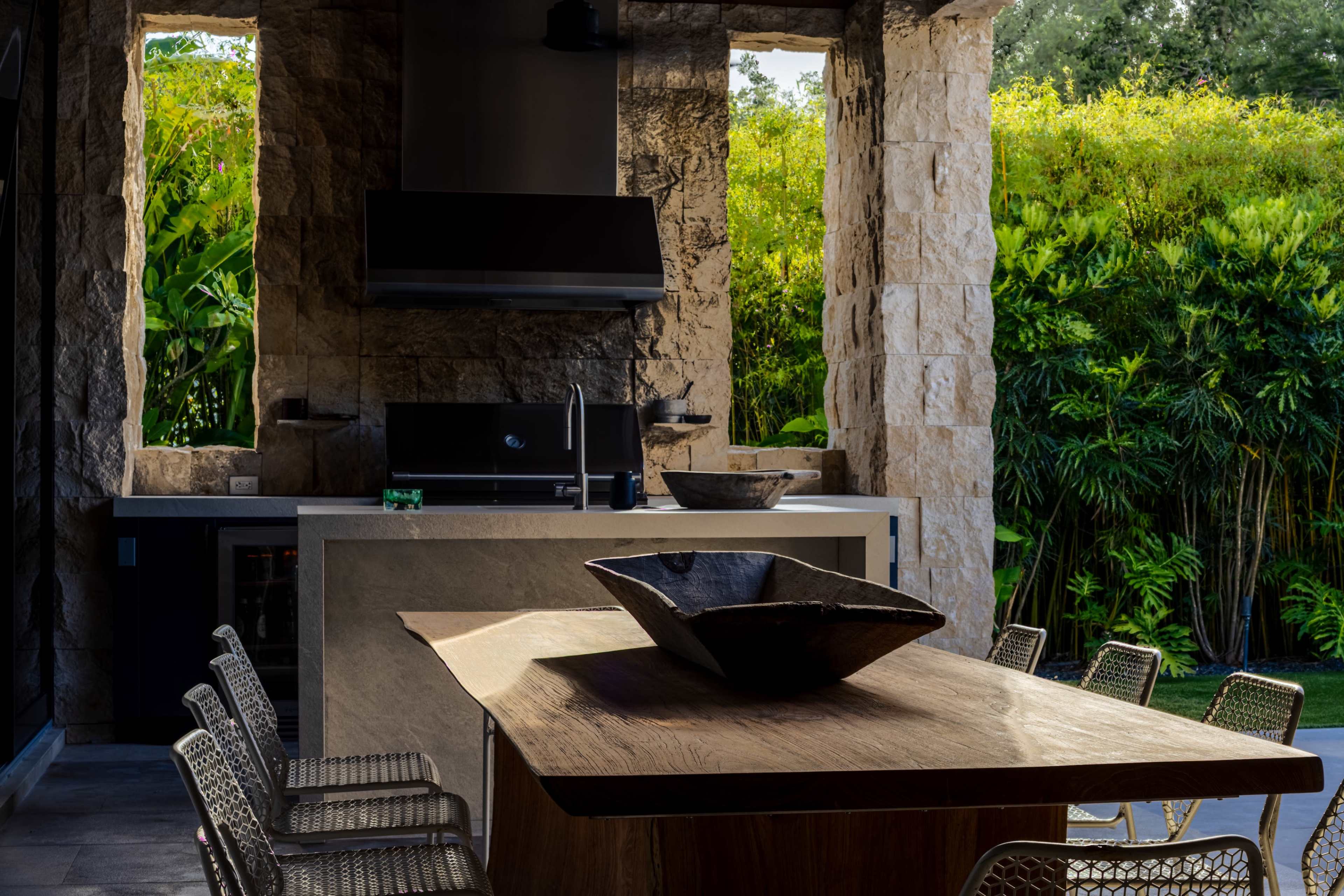 The image shows an outdoor kitchen area featuring a stone countertop, modern appliances, and a wooden dining table surrounded by metal chairs, with lush greenery in the background.