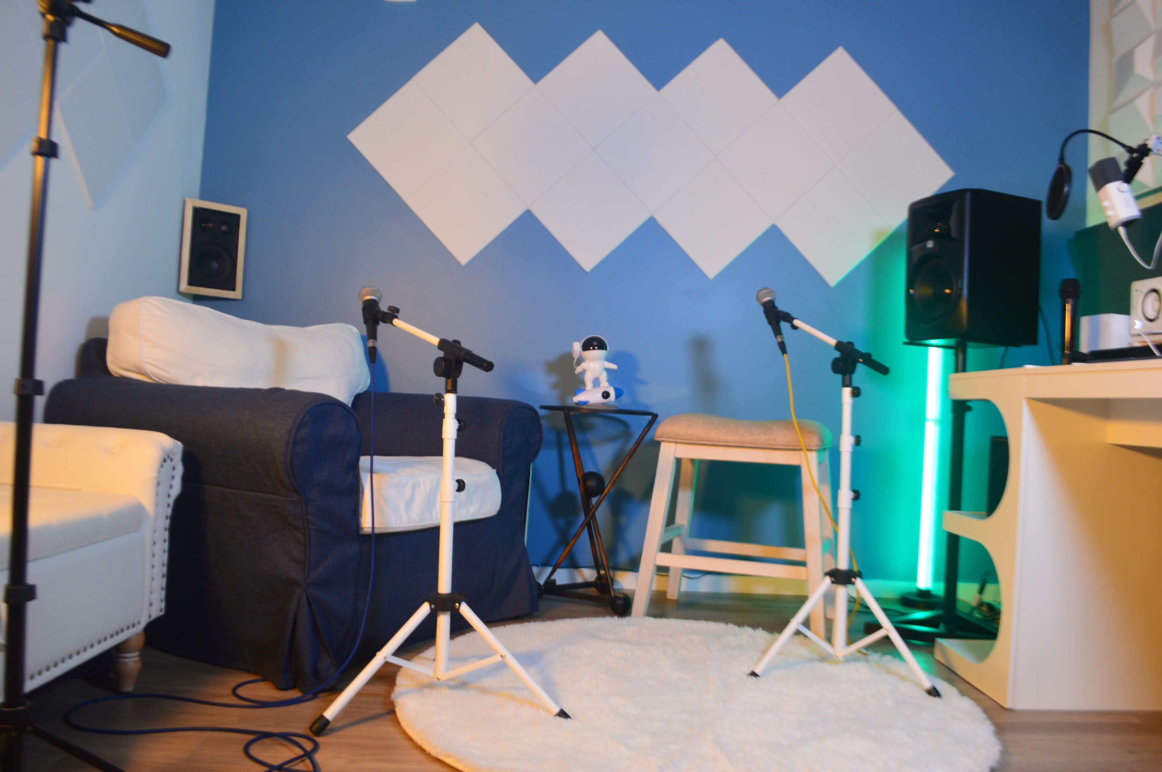 The image shows a small recording studio setup featuring a blue wall, a white armchair, two microphones on stands, a stool, and audio equipment.