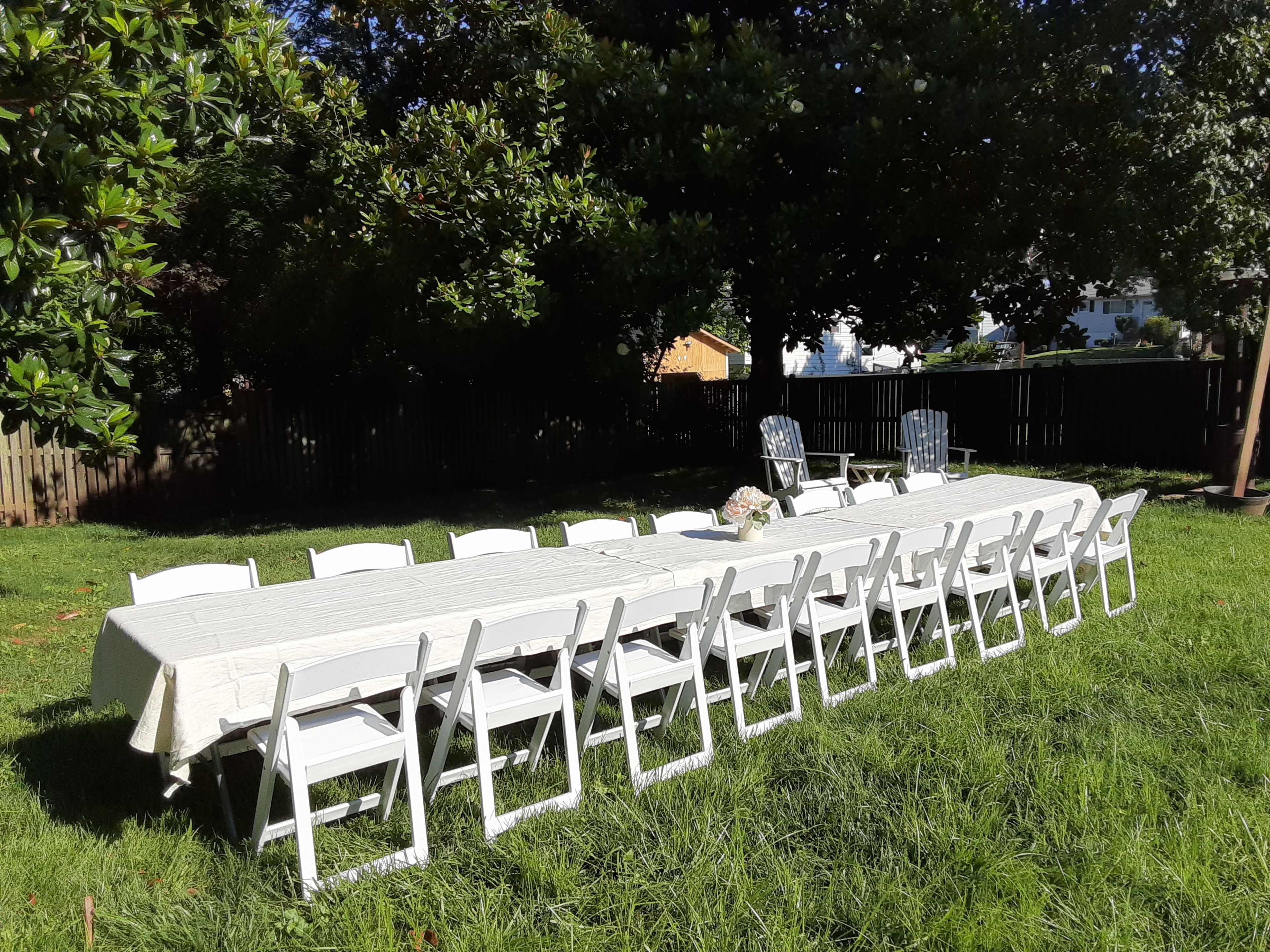 A long table set with white chairs is placed on green grass in a sunny backyard.