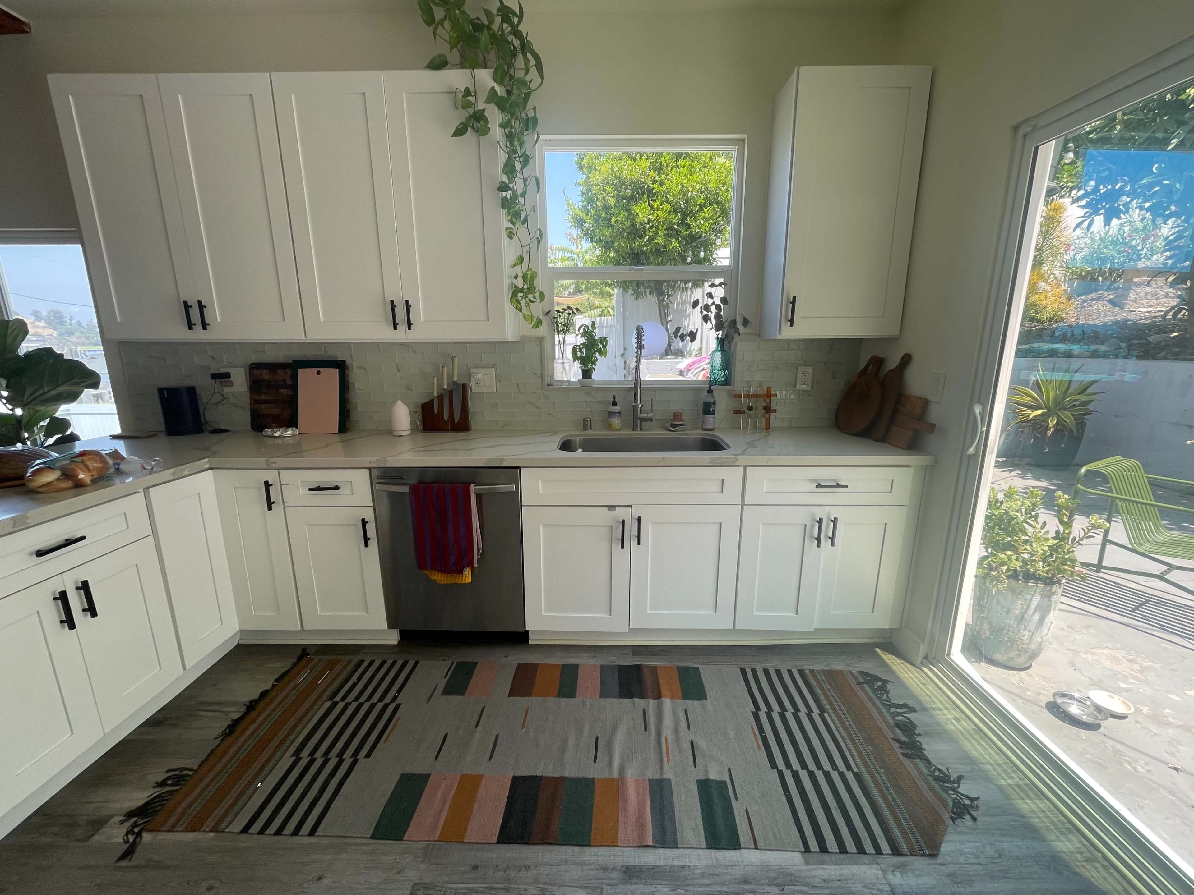 A bright kitchen features white cabinets, a stainless steel dishwasher, and a view of an outdoor area through a large sliding glass door.