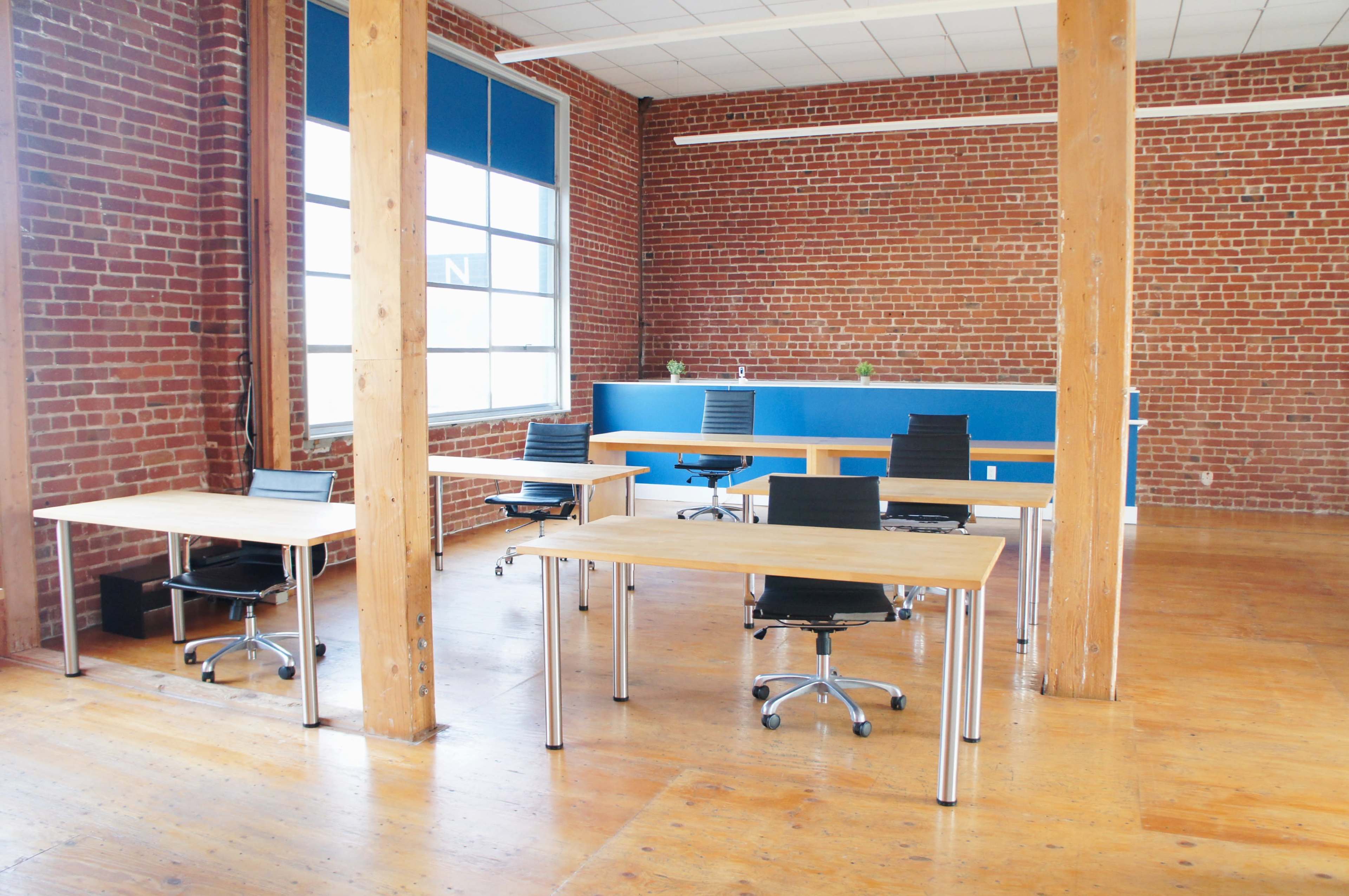 The image shows a spacious office with wooden flooring, brick walls, and several desks with rolling black chairs arranged around a central area.
