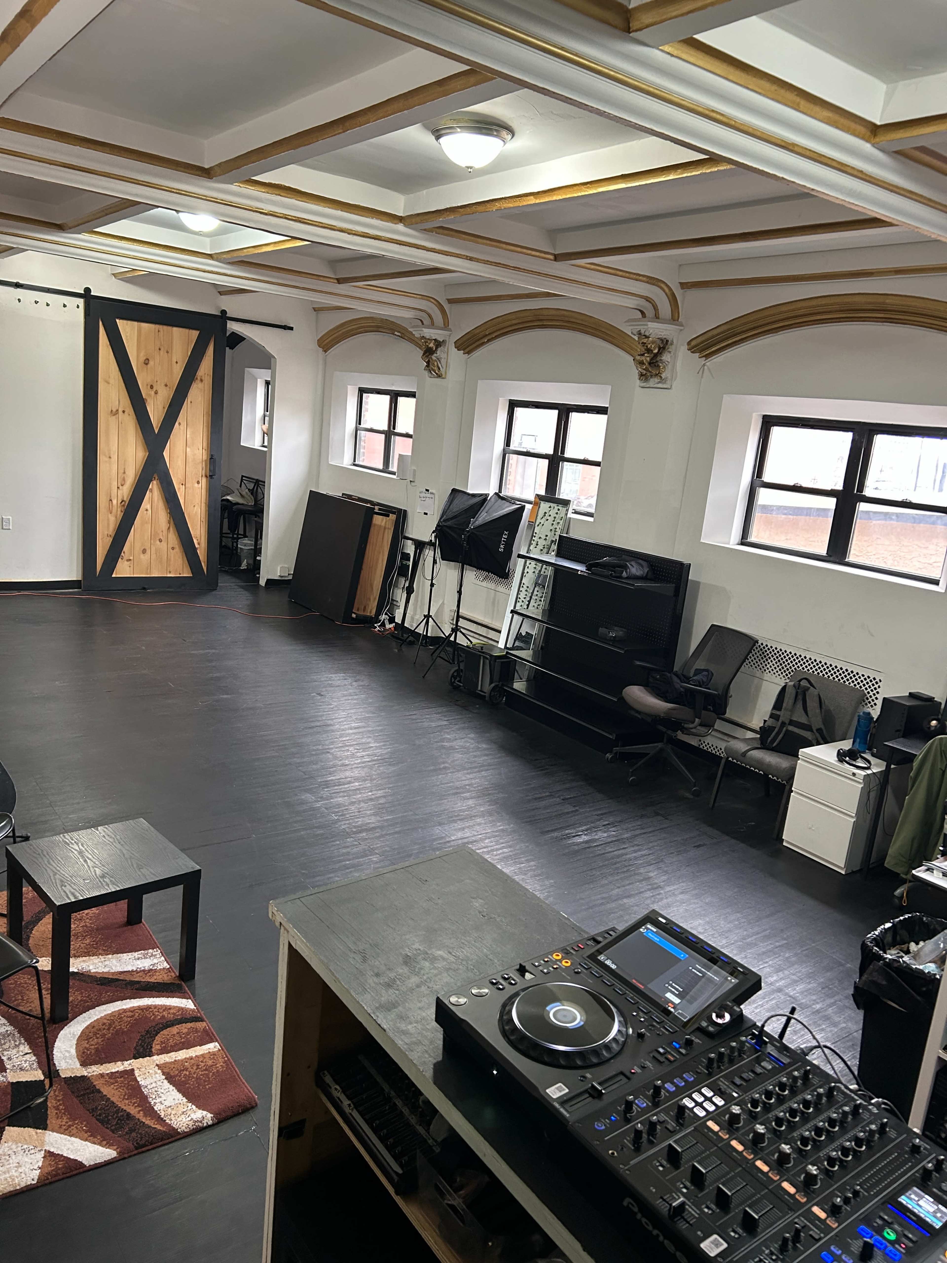The image depicts a spacious room with dark wooden flooring and large windows, featuring a DJ setup in the foreground, seating areas, and a sliding barn door.