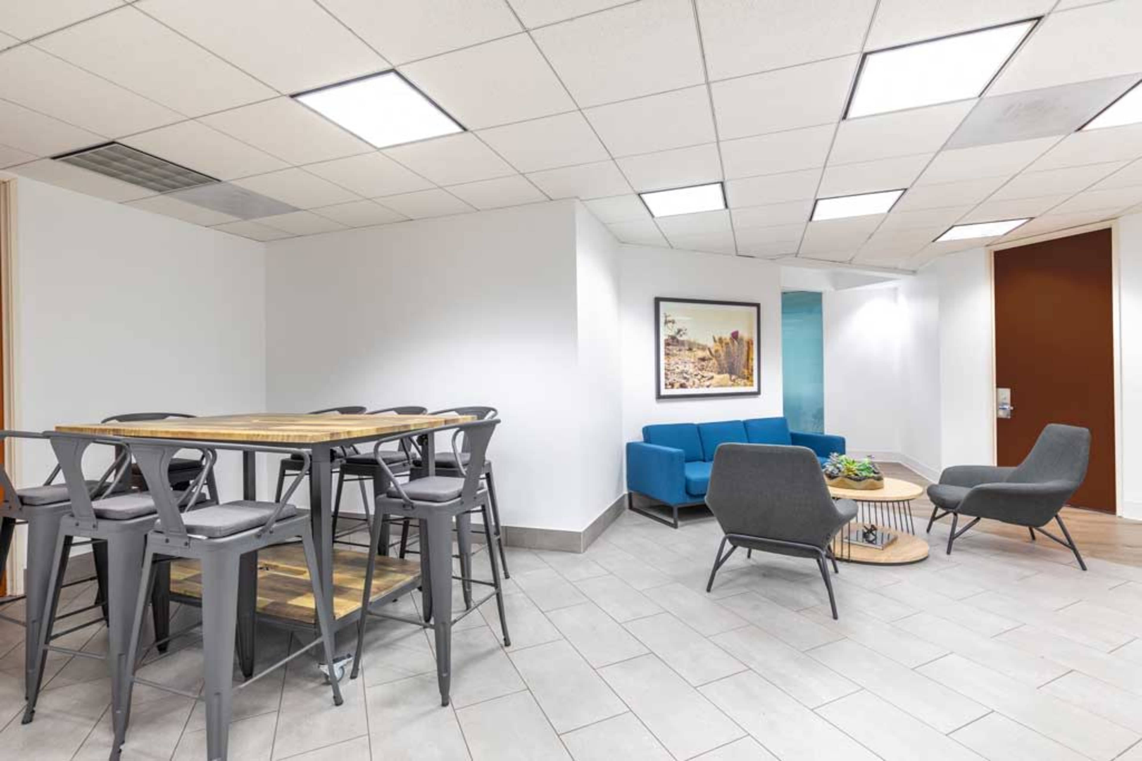 A modern office break area with a high table and metal stools, blue seating, and a small coffee table.