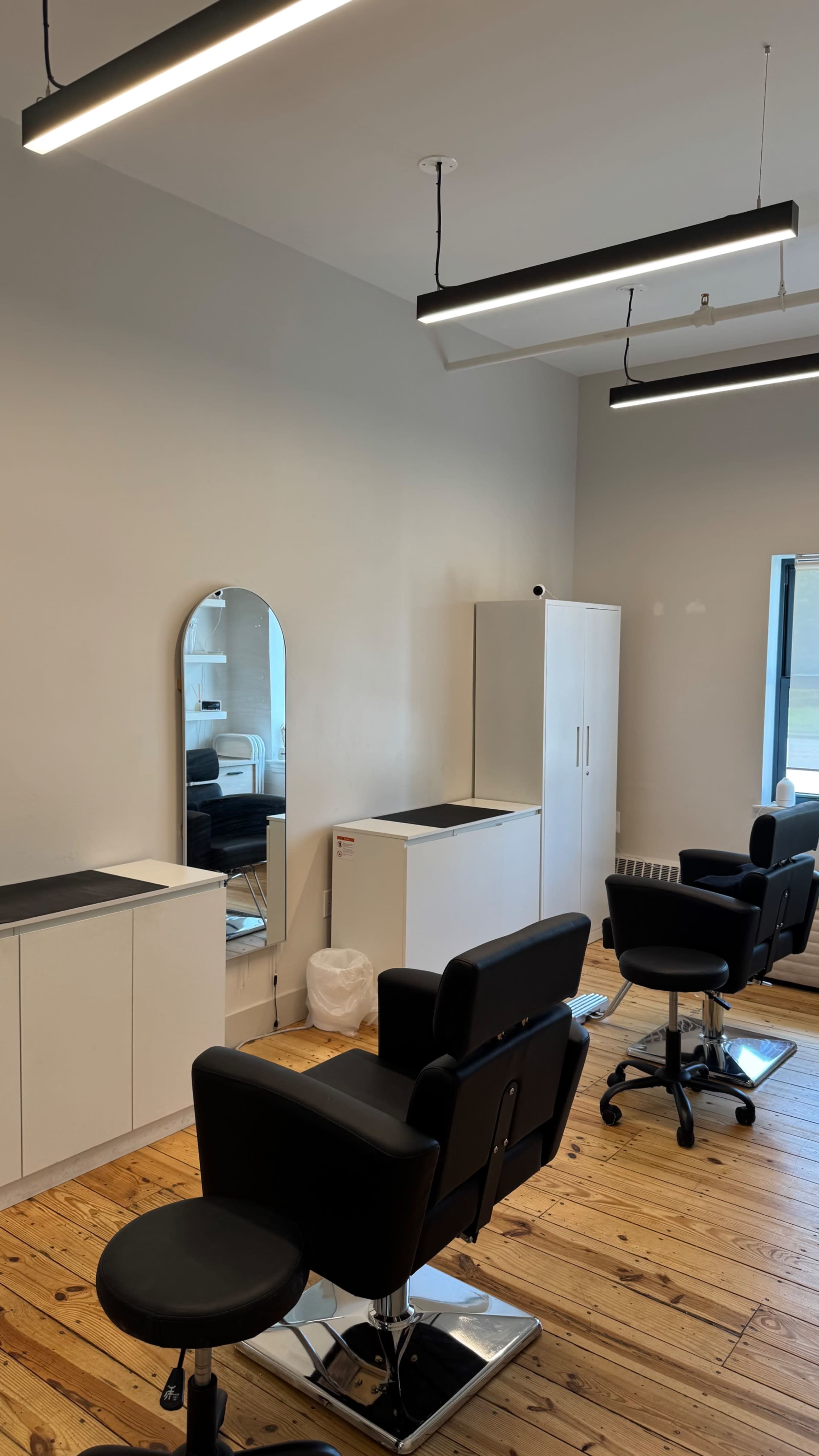 The image shows a modern hair salon featuring two black salon chairs, a large mirror on the wall, and white cabinetry against a light-colored wall.