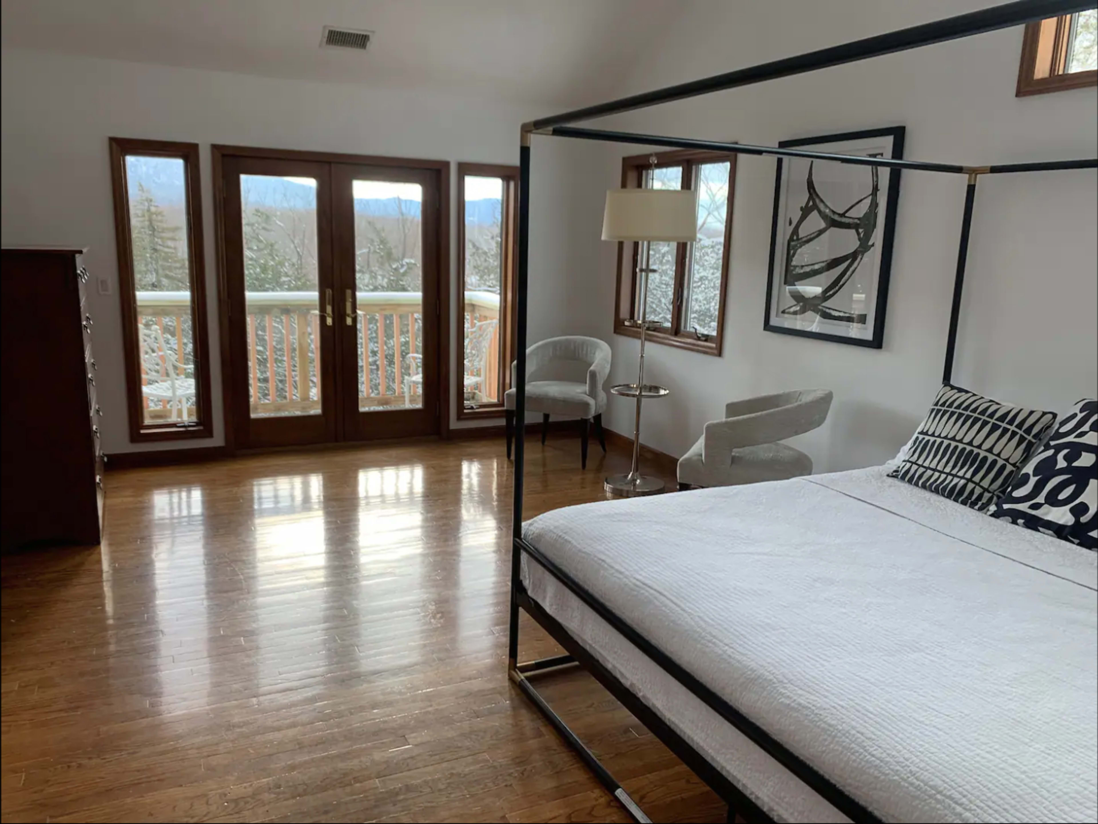 A spacious bedroom featuring a four-poster bed, a seating area, large windows with a view, and hardwood flooring.