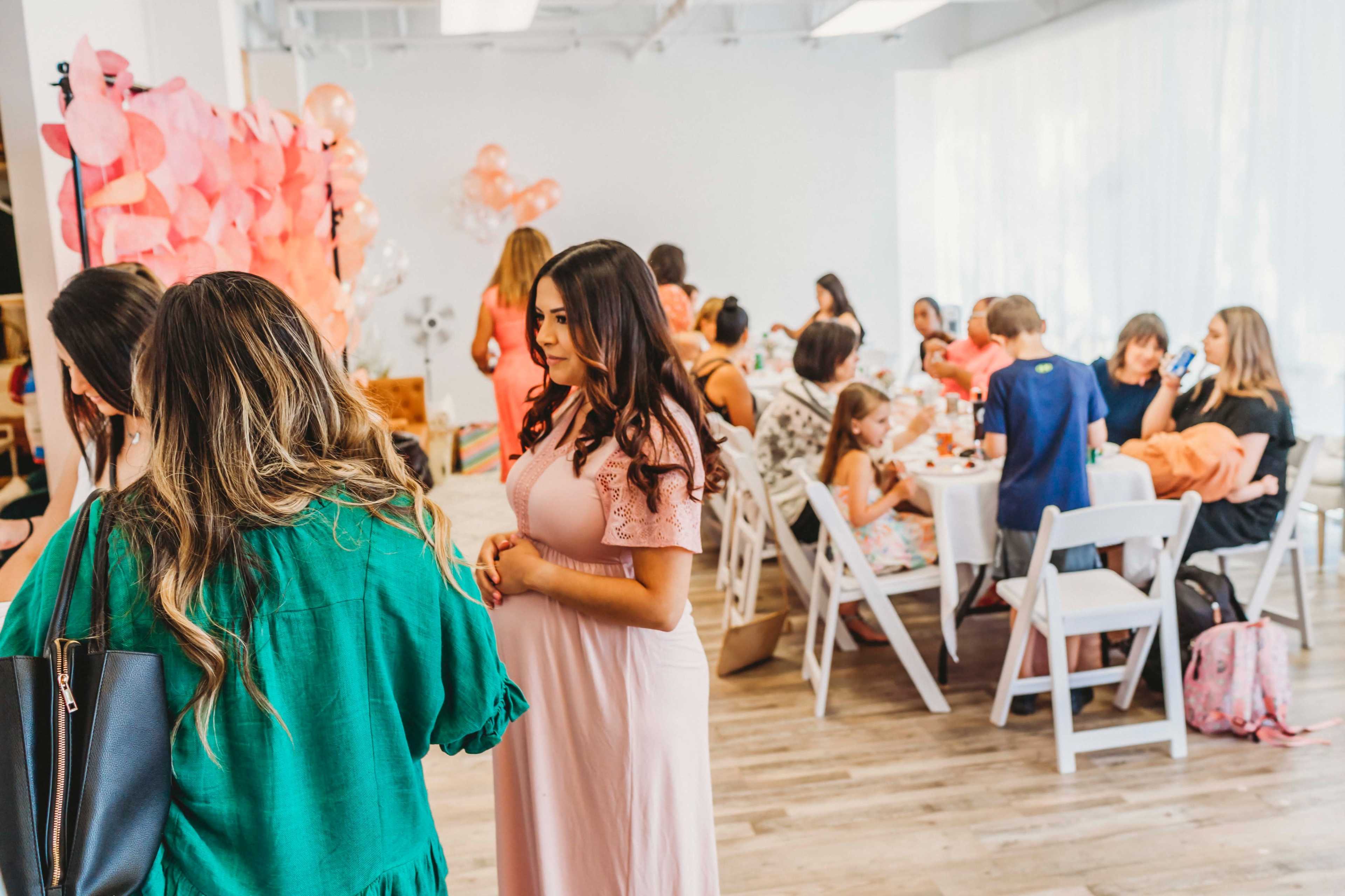 A group of adults and children are gathered in a bright, spacious room decorated with pink accents, engaged in conversation and activities at tables.