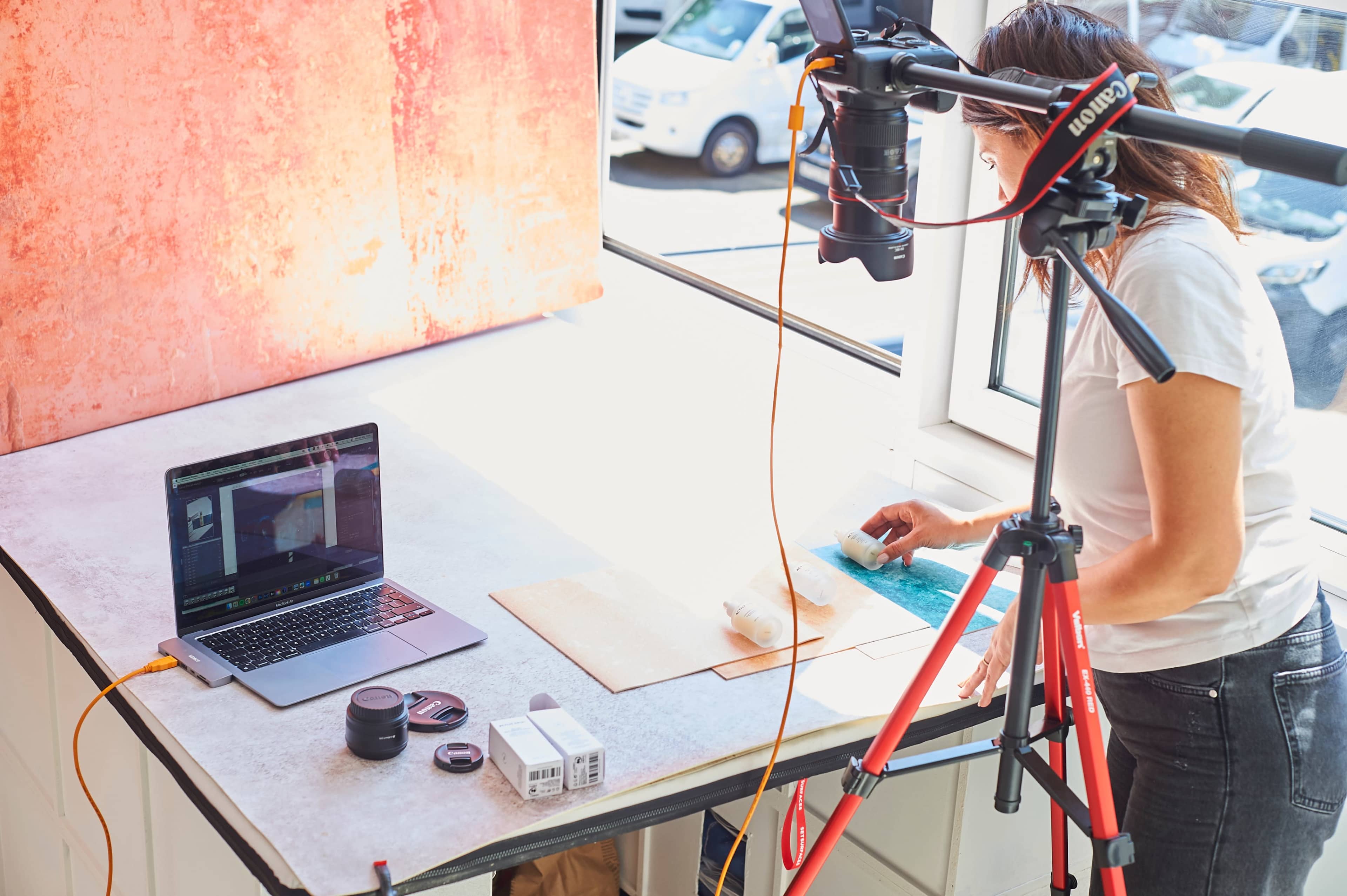 A person is preparing materials on a table for a photography shoot, with a camera mounted on a tripod and a laptop displaying software.
