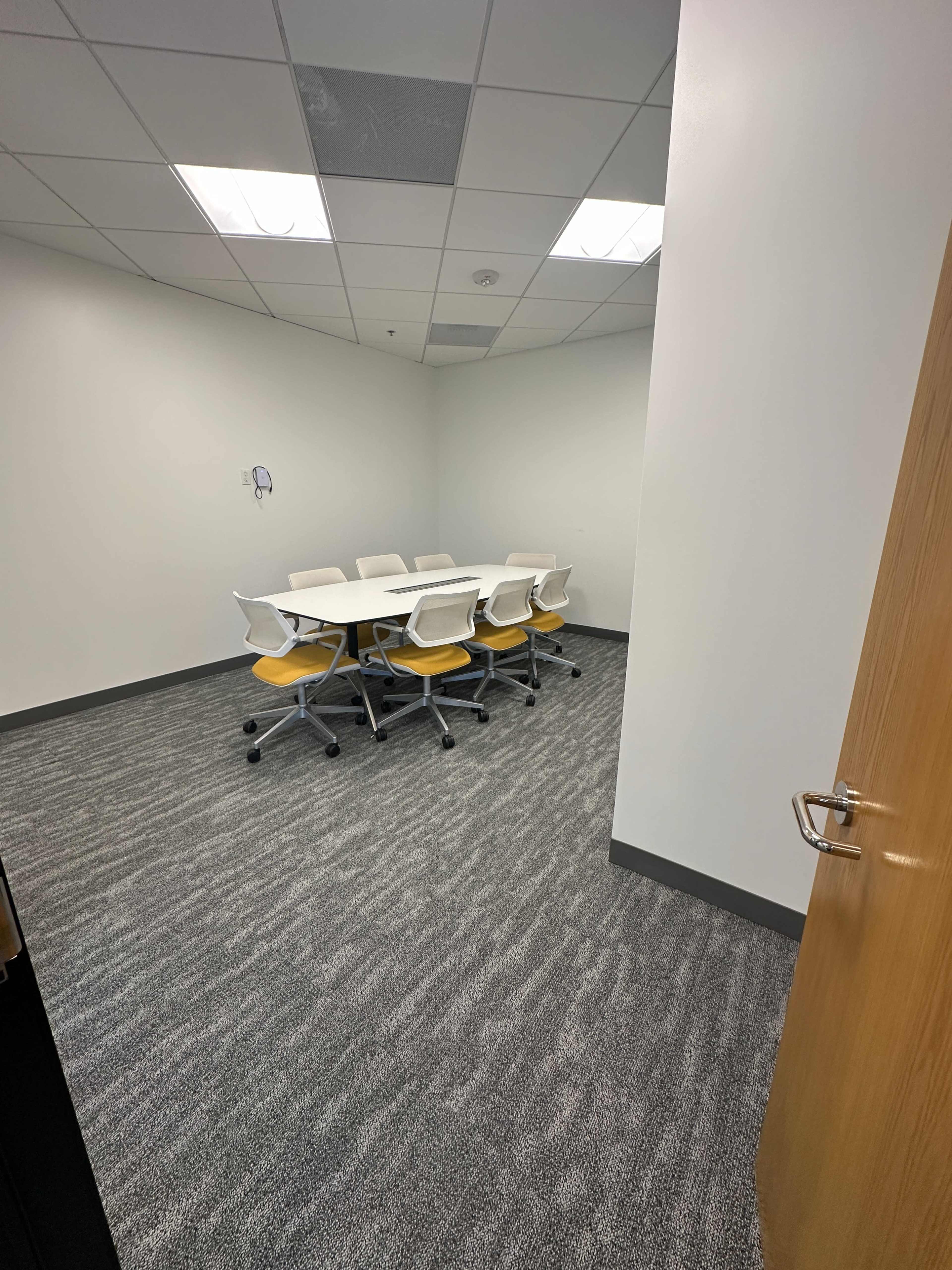 The image shows a small, empty conference room featuring a white table surrounded by eight chairs with yellow cushions, and a door partially open at the entrance.