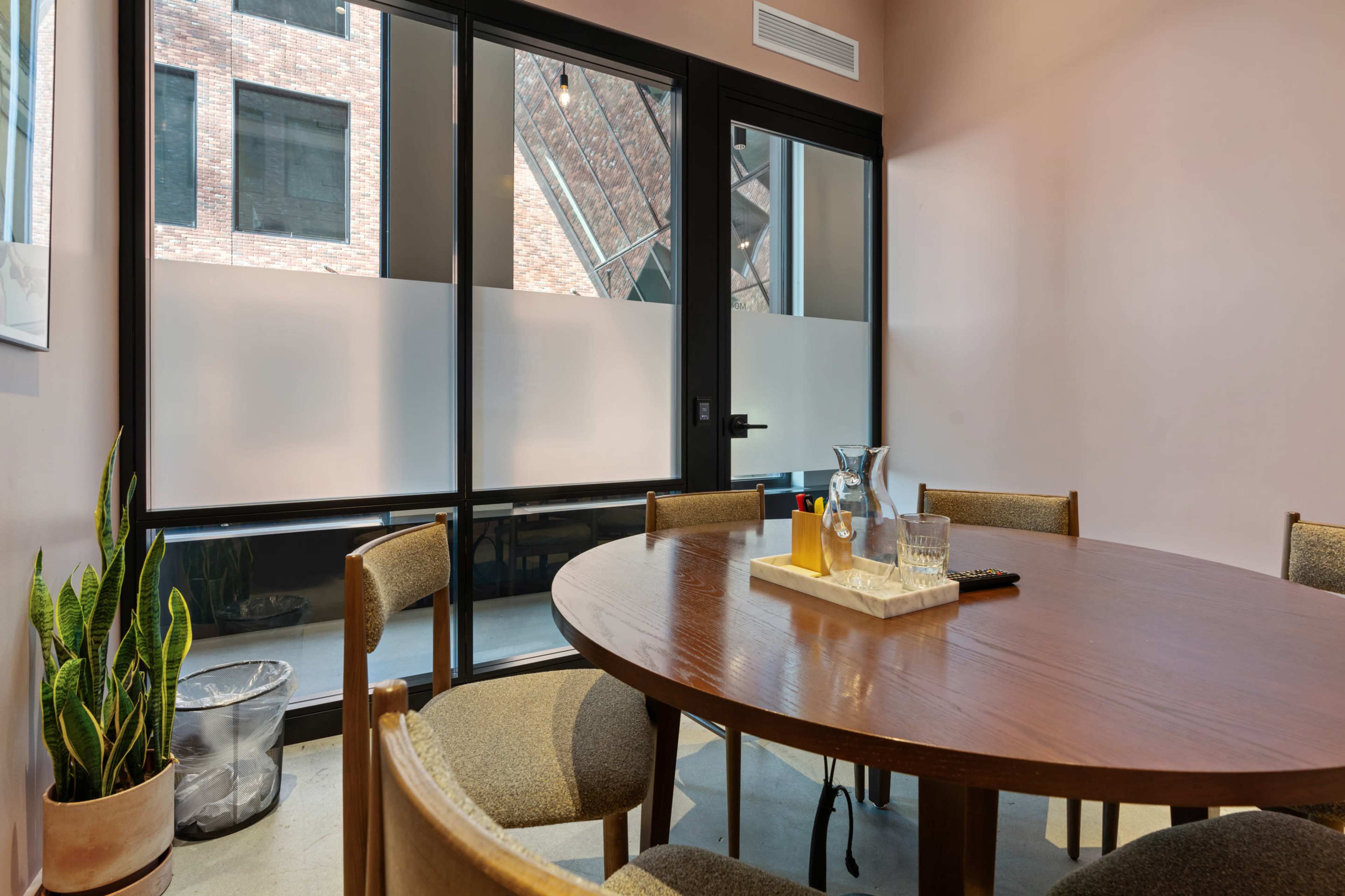 A wooden conference table is set with a pitcher and glasses, while a large window offers a view of the exterior brick wall.