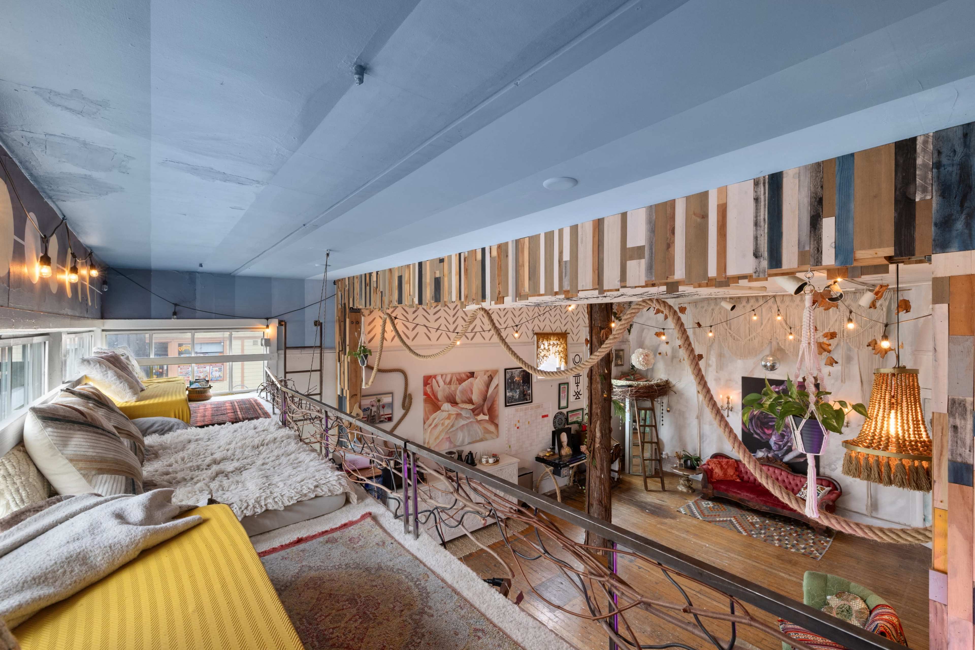 The image shows a brightly lit, artistic loft space with a cozy seating area on a raised balcony overlooking a colorful, decoratively arranged living area below.
