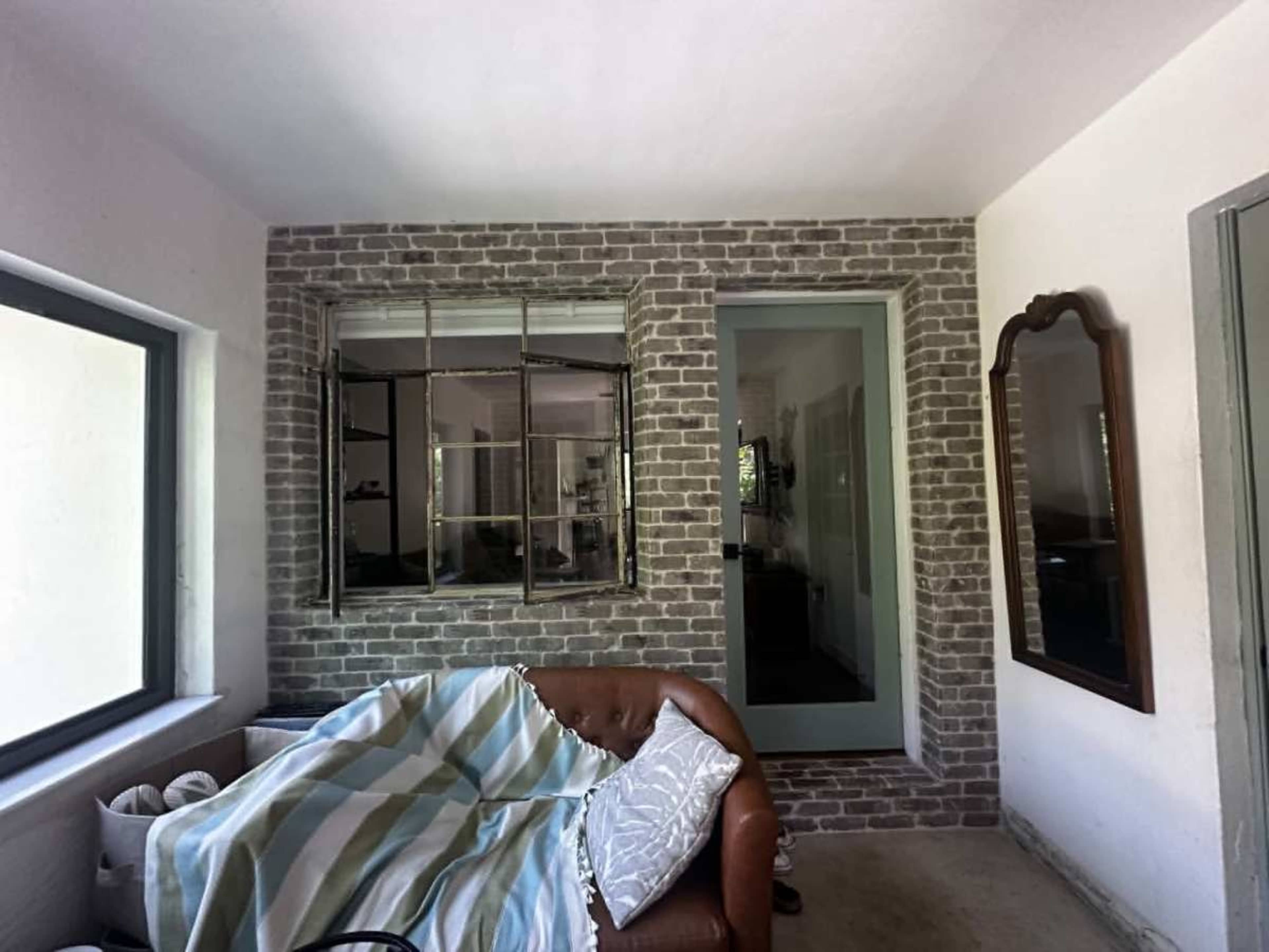 The image shows a room with exposed brick walls, a large window, a brown leather couch draped with a striped blanket, and a mirror mounted on the wall.