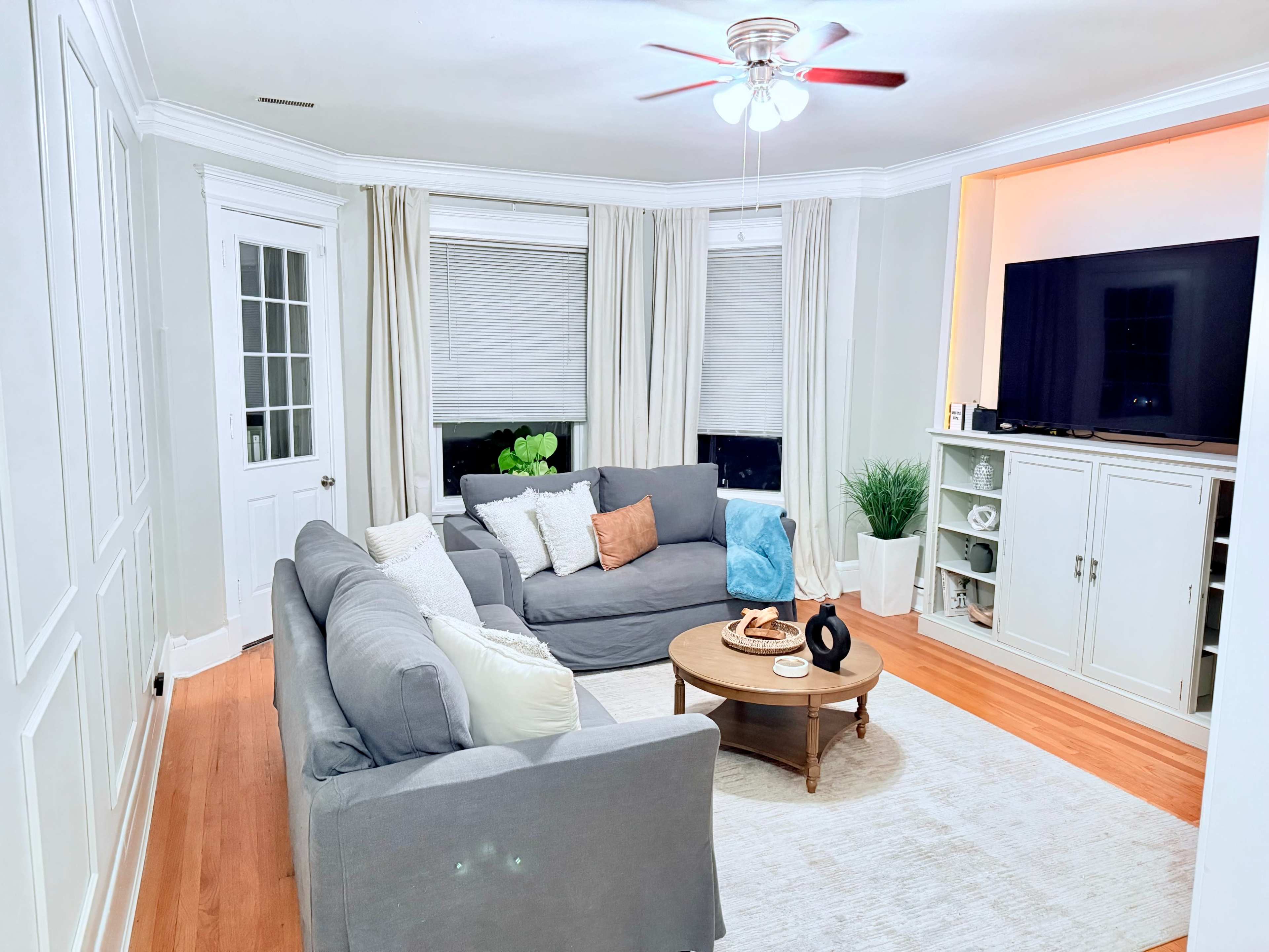 The living room features a gray sectional sofa, a round coffee table, and a light-colored area rug, with a television mounted on the wall and closed cabinetry in the corner.
