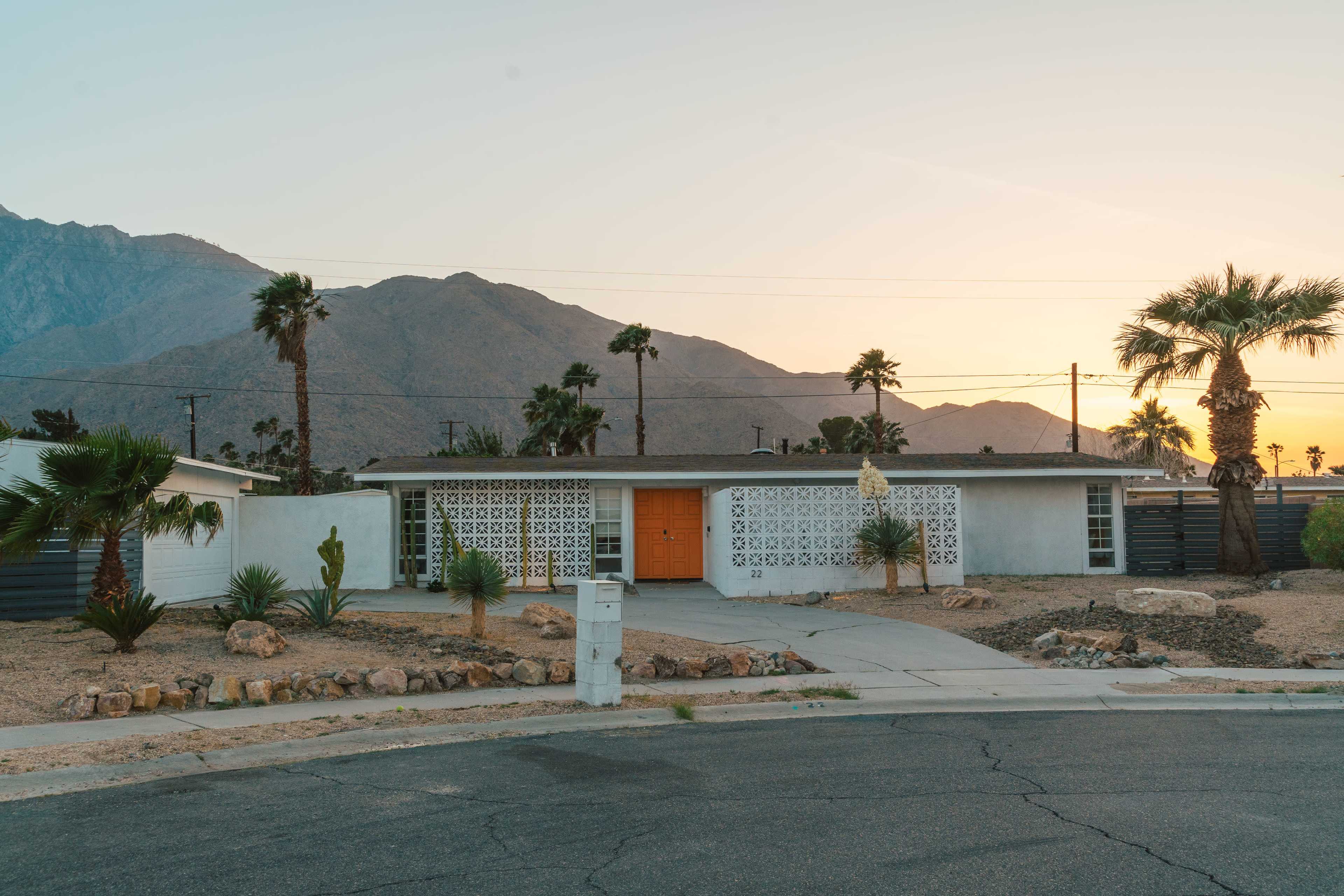 A modern house with a patterned concrete wall is situated at the corner of a street, with mountains and palm trees in the background during sunset.