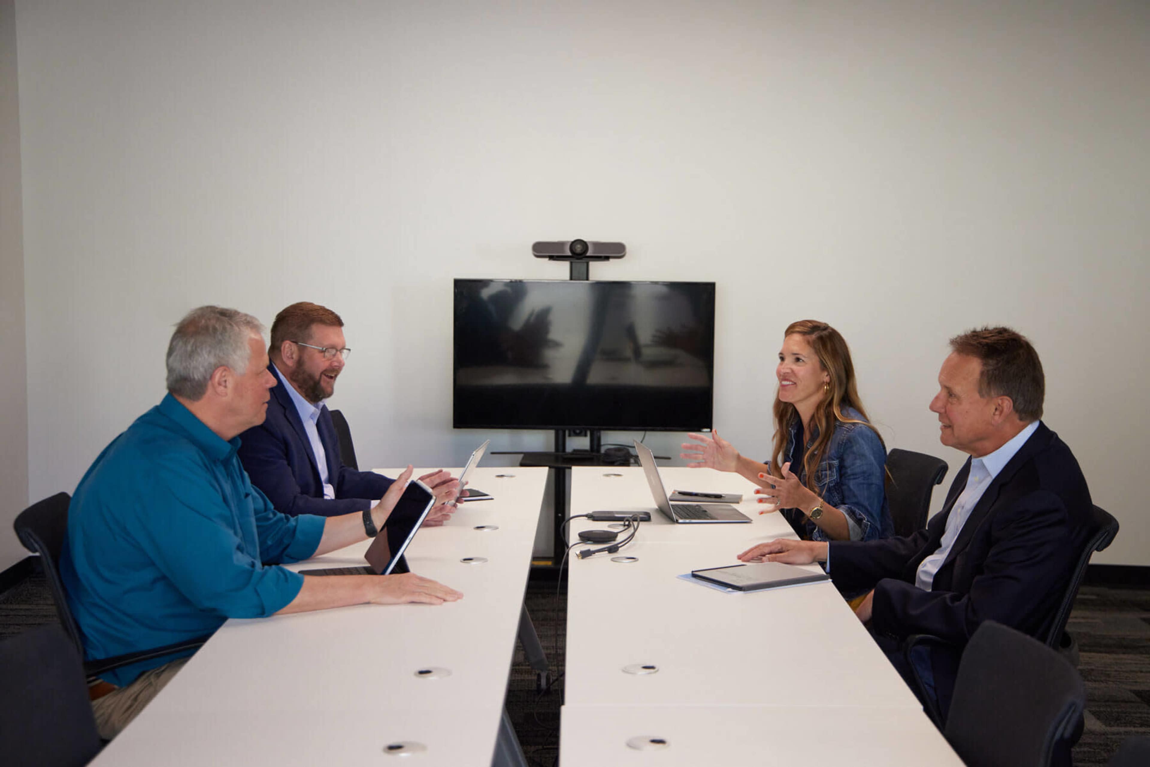 Five professionals are seated around a long white conference table engaged in a discussion, with a television monitor displaying a blurred image in the background.