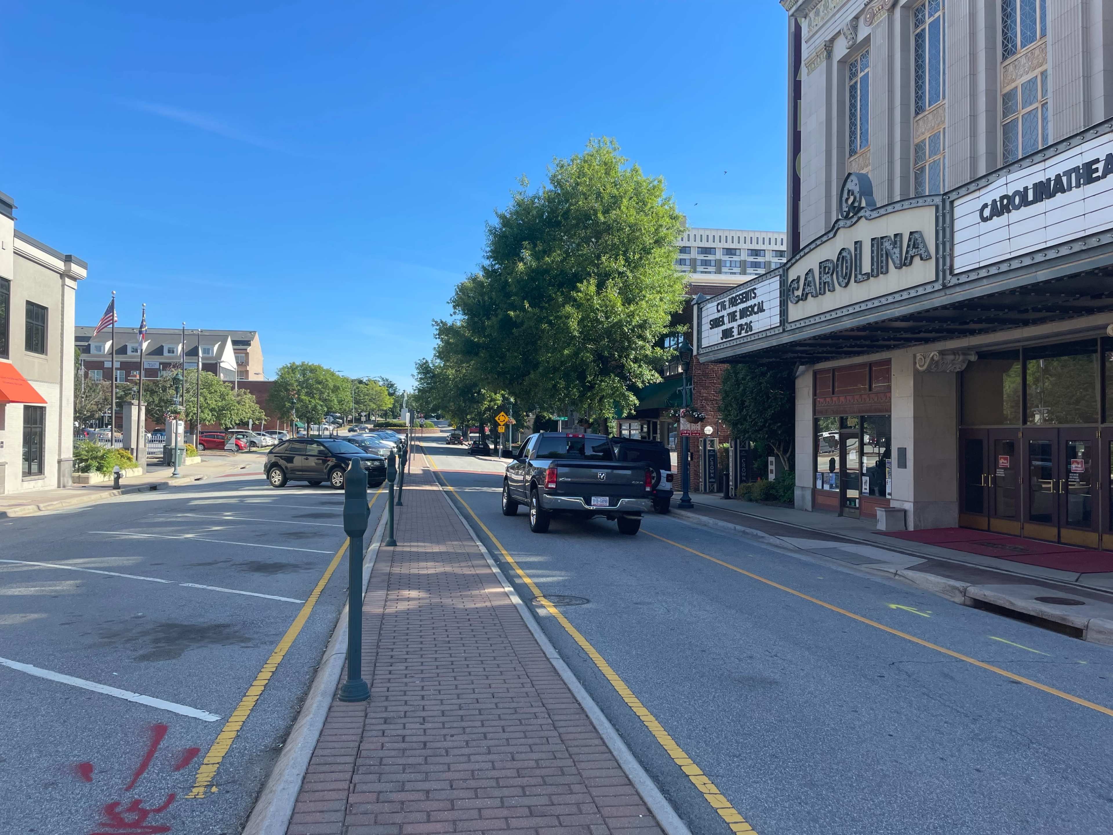 A black pickup truck drives down a tree-lined street adjacent to a theater marked "CAROLINA."