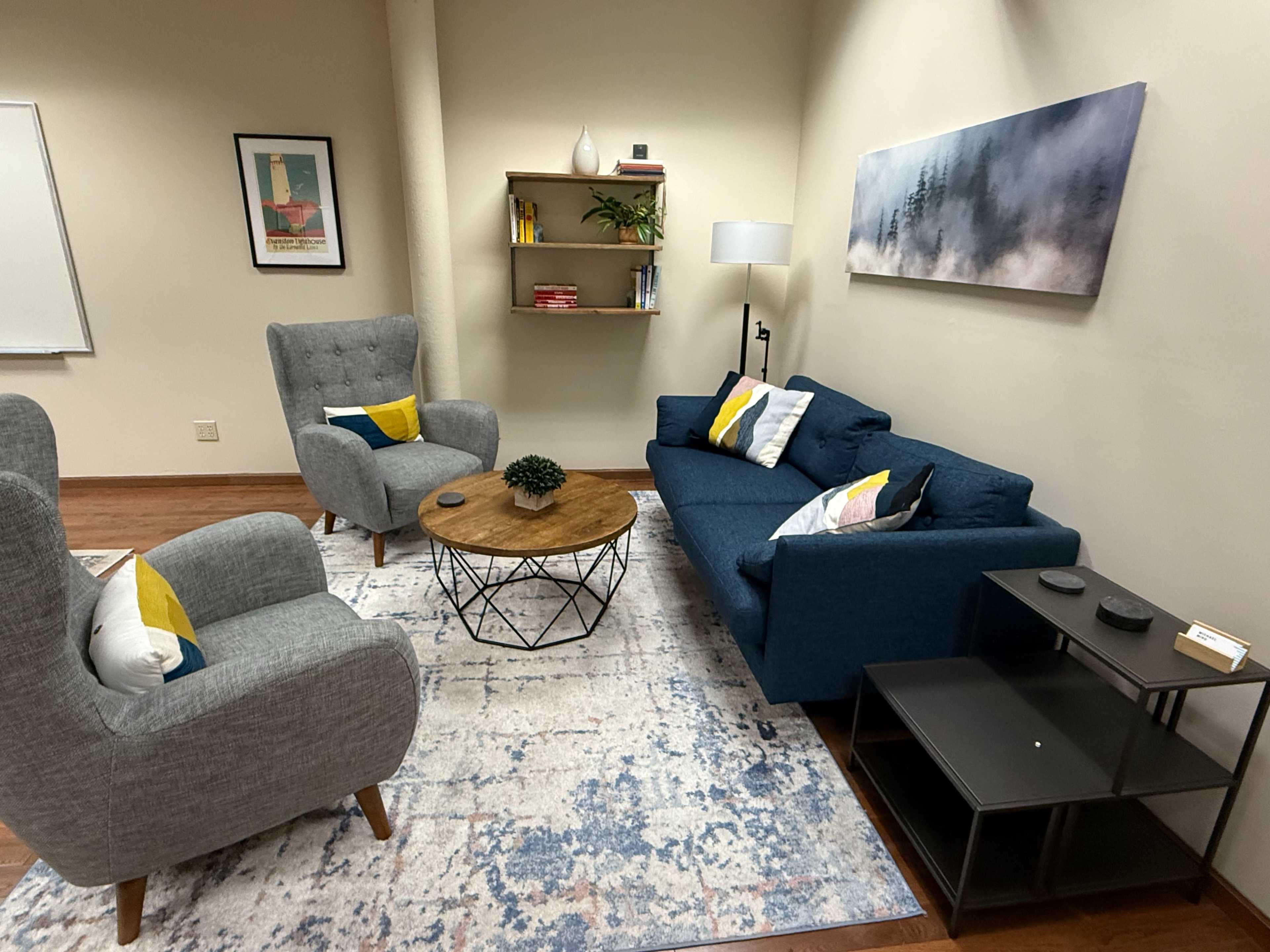 The image shows a cozy lounge area with two armchairs and a sofa arranged around a wooden coffee table, accompanied by a small bookshelf and a floor lamp against a neutral-colored wall.