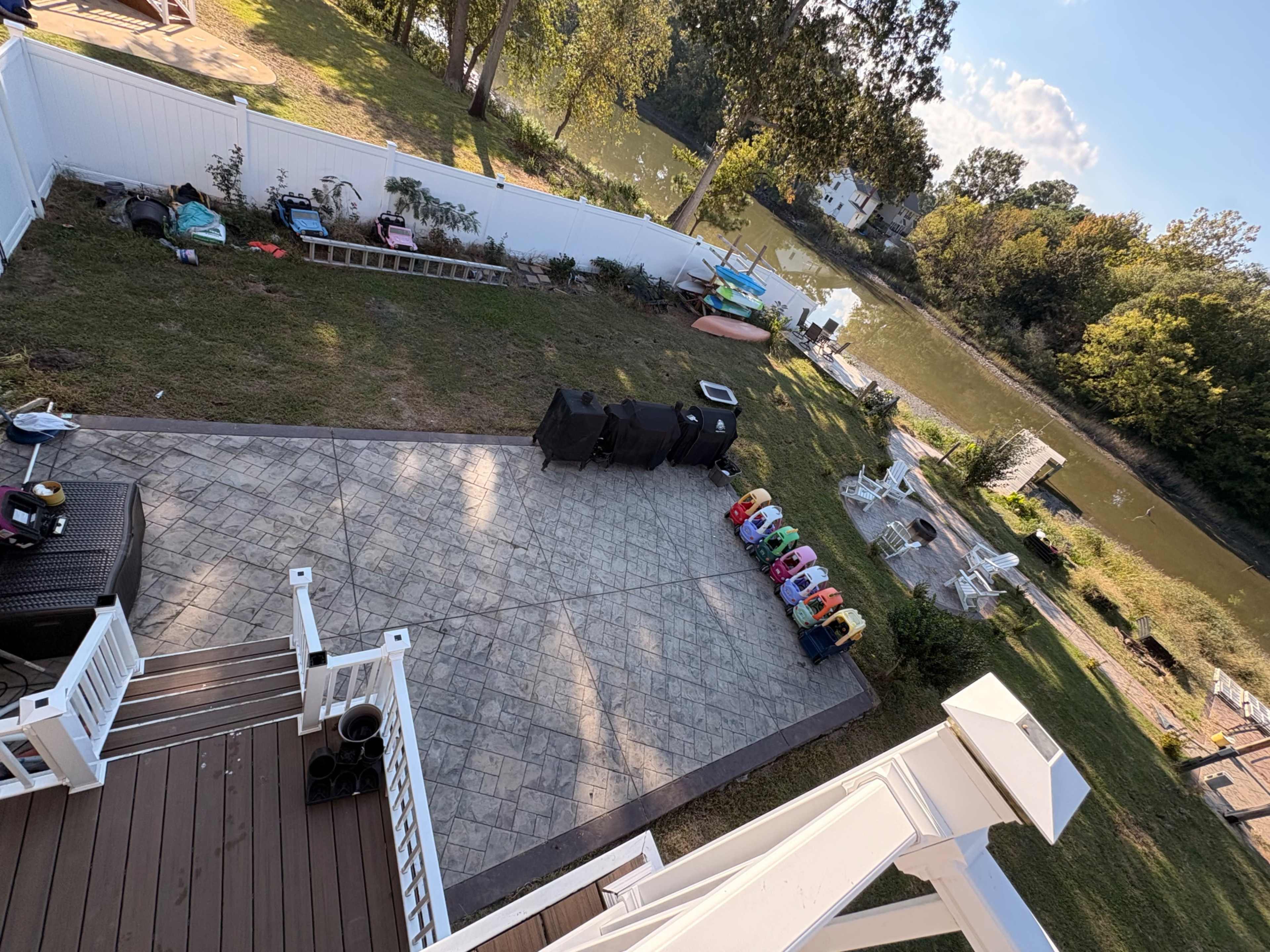 The image shows a backyard area with a stone patio, garden, and a river in the background, featuring various items such as a ladder, garbage bins, and colorful children's ride-on toys.