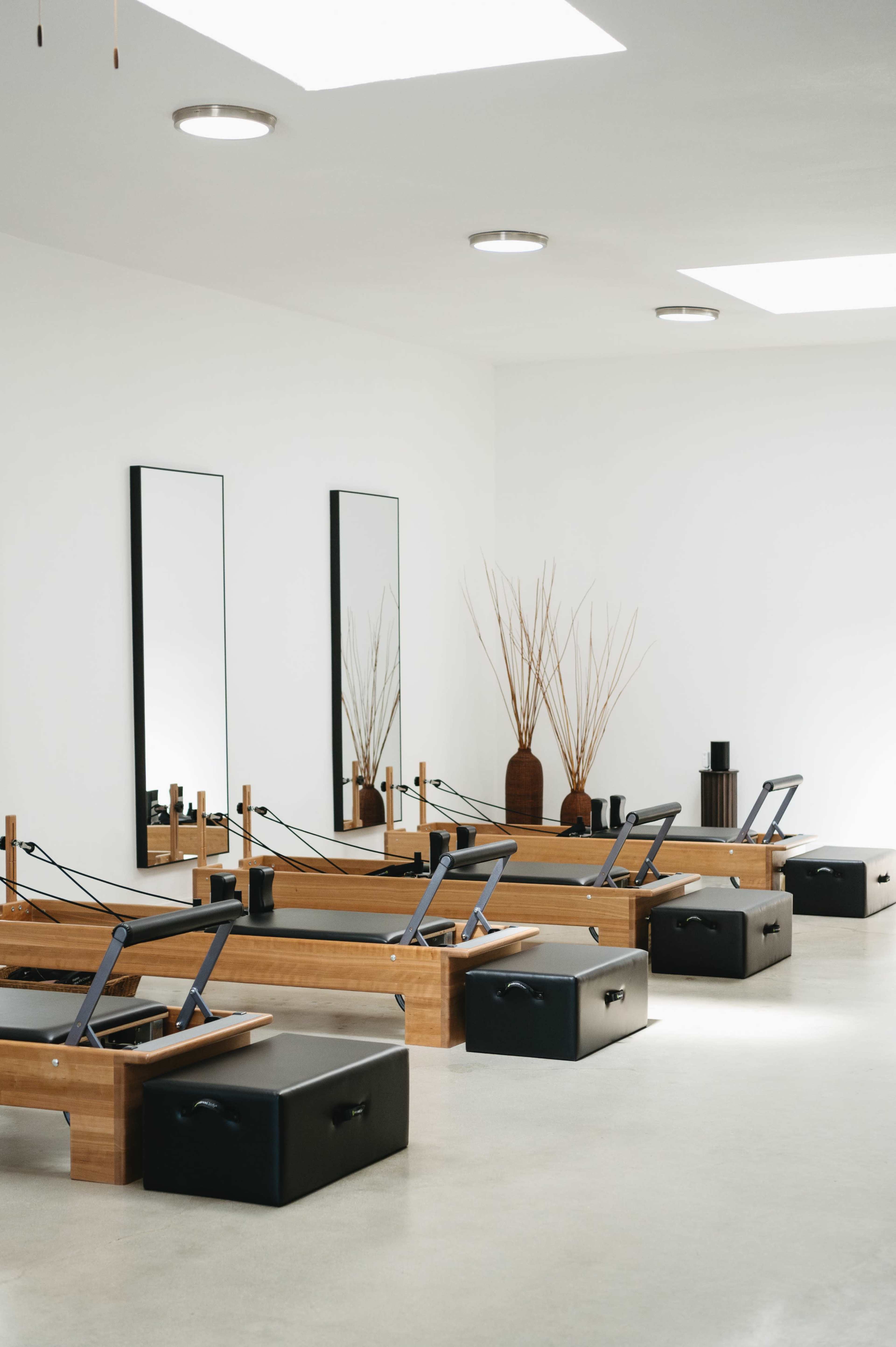 The image shows a spacious pilates studio with wooden reformer equipment and large mirrors against a white wall.