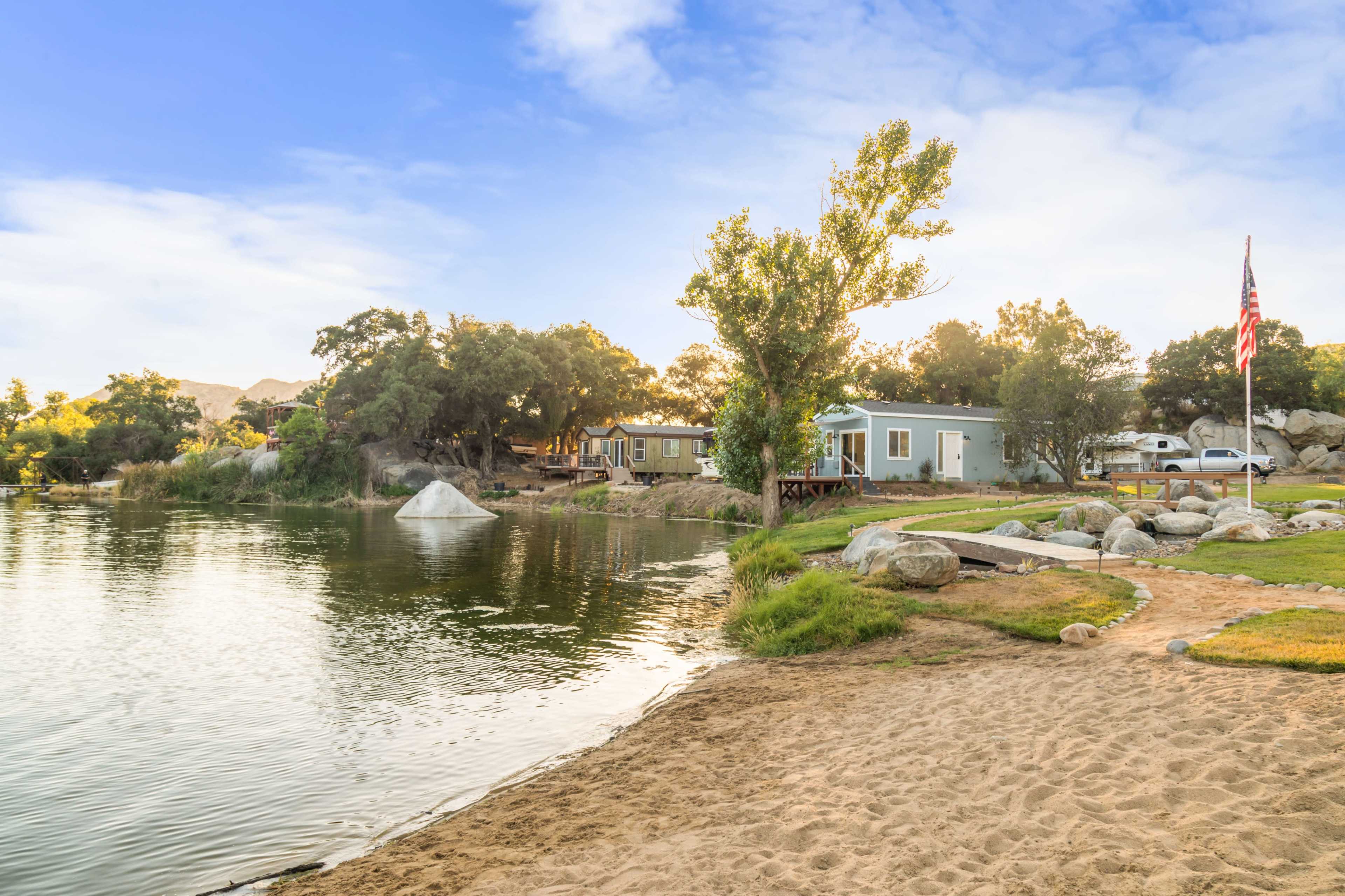 The image shows a serene lakeside scene with a sandy shore, surrounded by rocks, trees, and a few cabins.