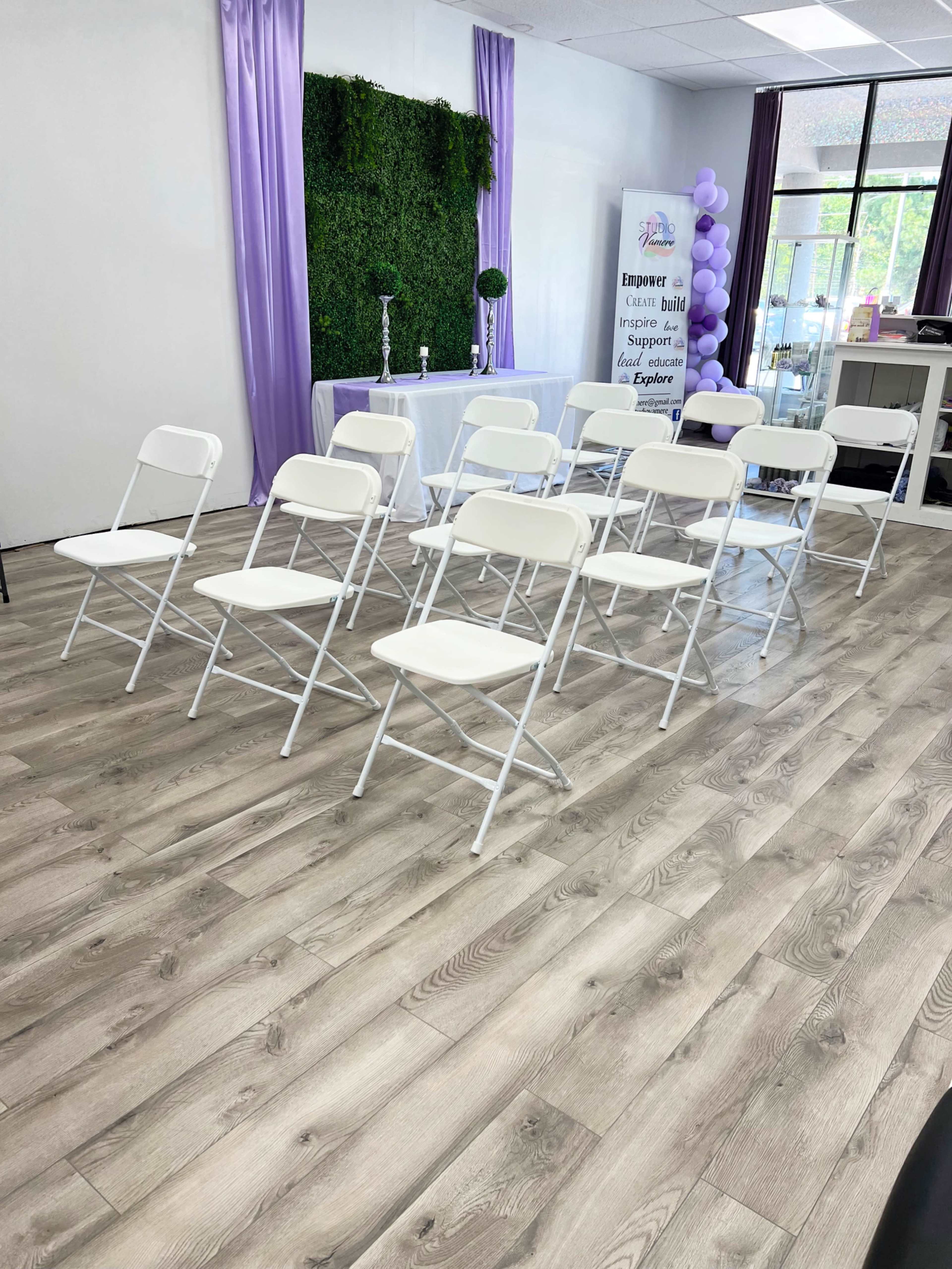 A row of white folding chairs is set up in a room with a green wall and decorative elements, including purple accents and a table at the front.