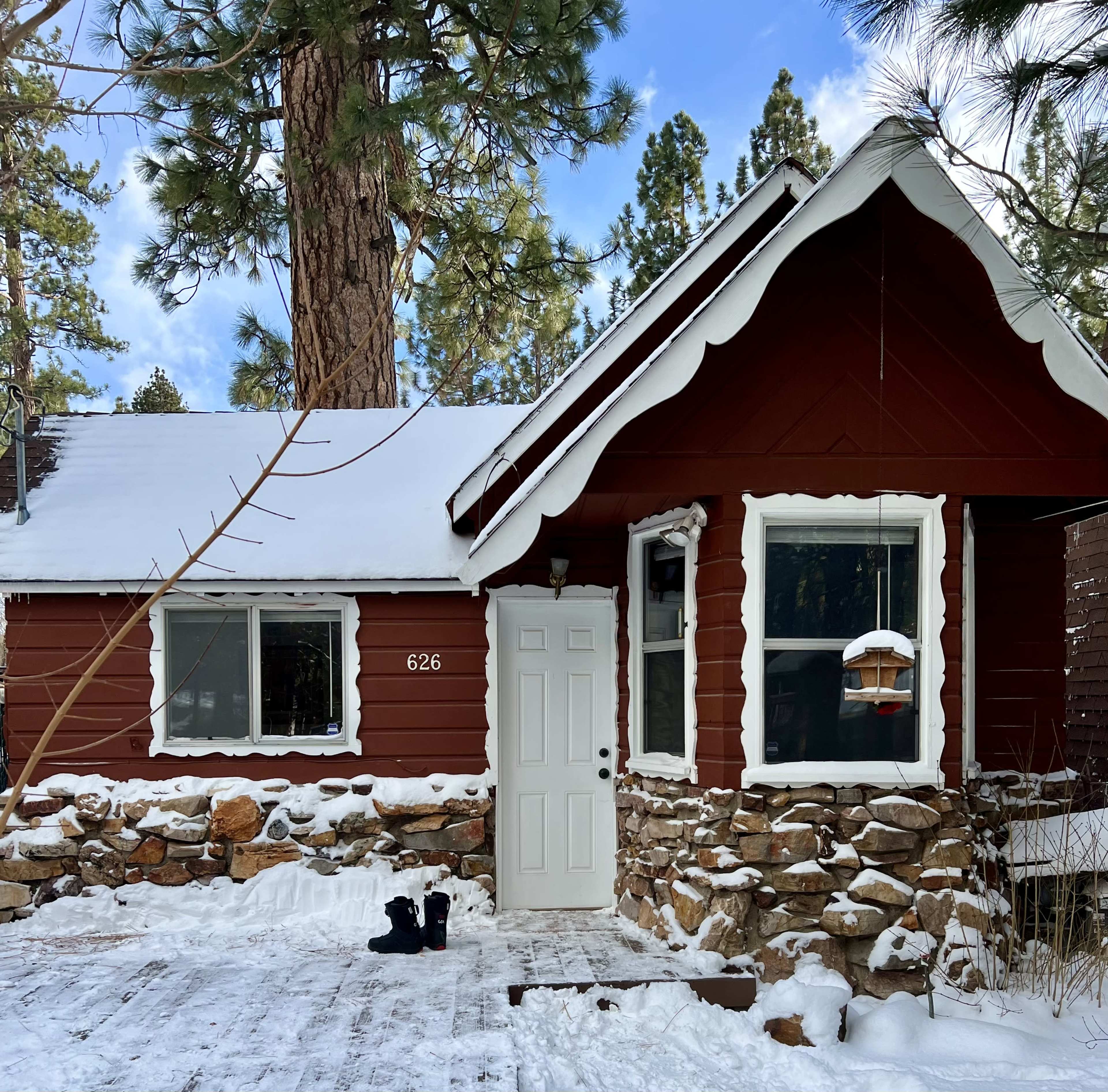 A small brown cabin with a stone foundation is surrounded by snow and pine trees, featuring a front door and a wooden porch.